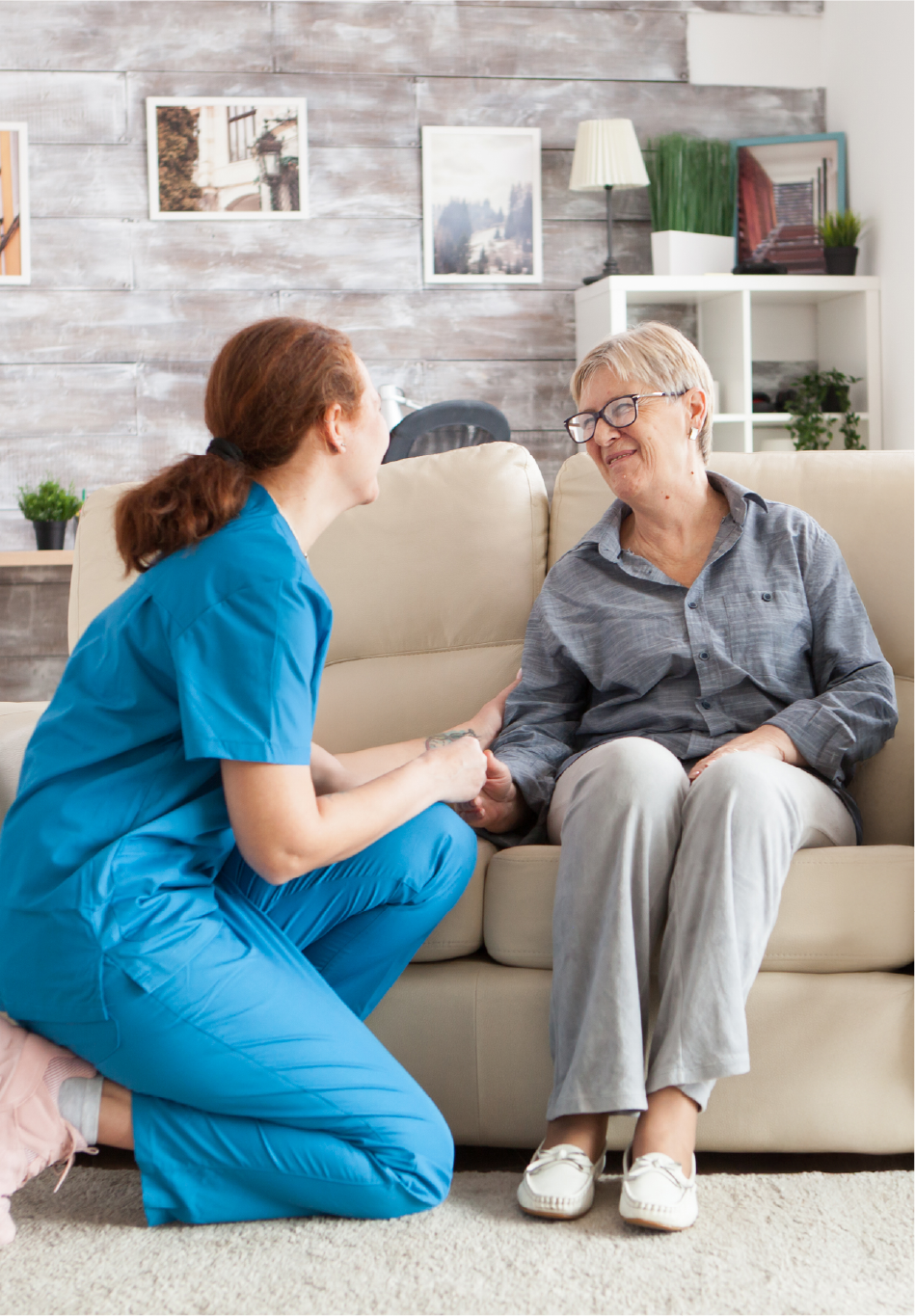 A caregiver in blue scrubs kneeling and holding hands with an elderly woman with short gray hair and glasses, seated on a cream-colored sofa in a cozy room with wooden wall paneling and framed pictures.