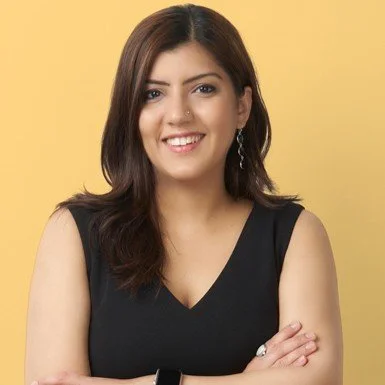 A woman with shoulder-length dark brown hair, wearing a black sleeveless top, smiling with her arms crossed against a yellow background.