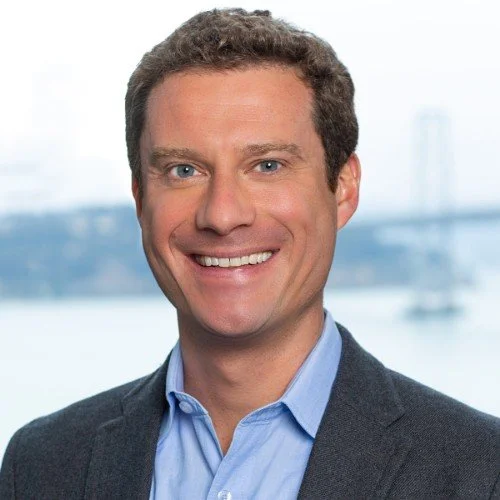 A smiling man with short brown hair wearing a blue dress shirt and dark blazer, with a blurred background of a bridge and water.