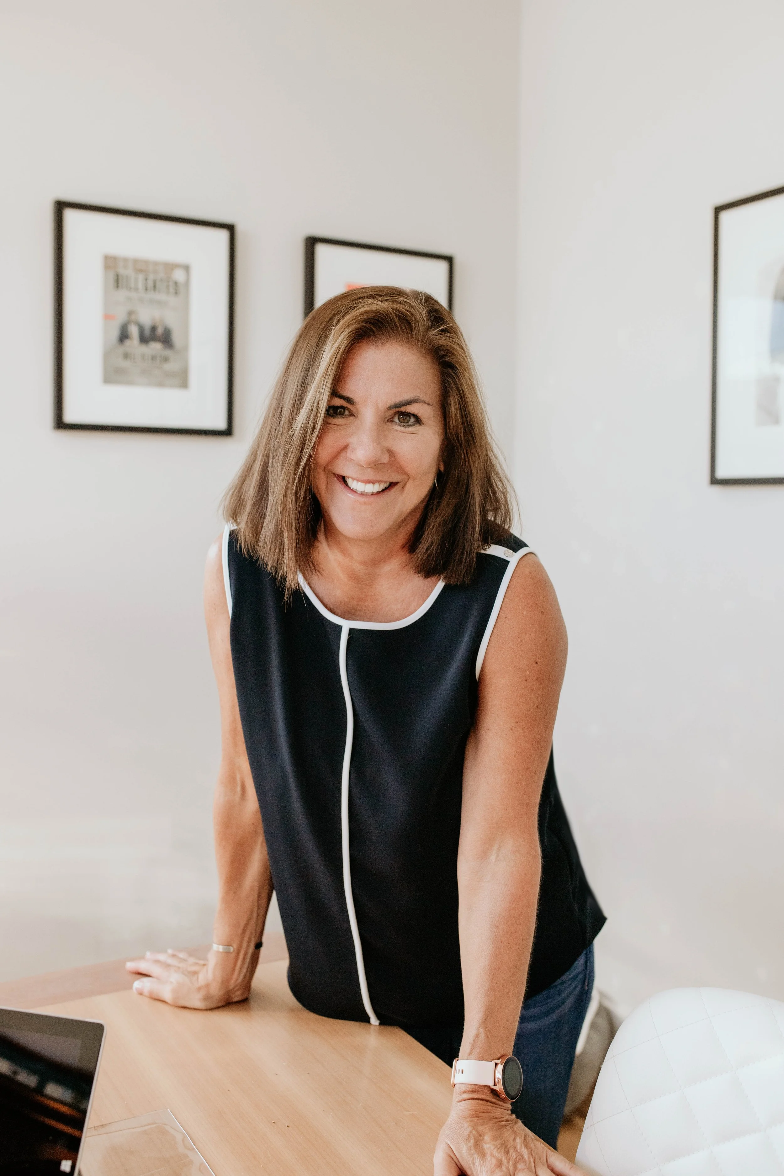 A woman with shoulder-length brown hair, wearing a sleeveless black top with white trim, leaning on a wooden table, smiling at the camera.