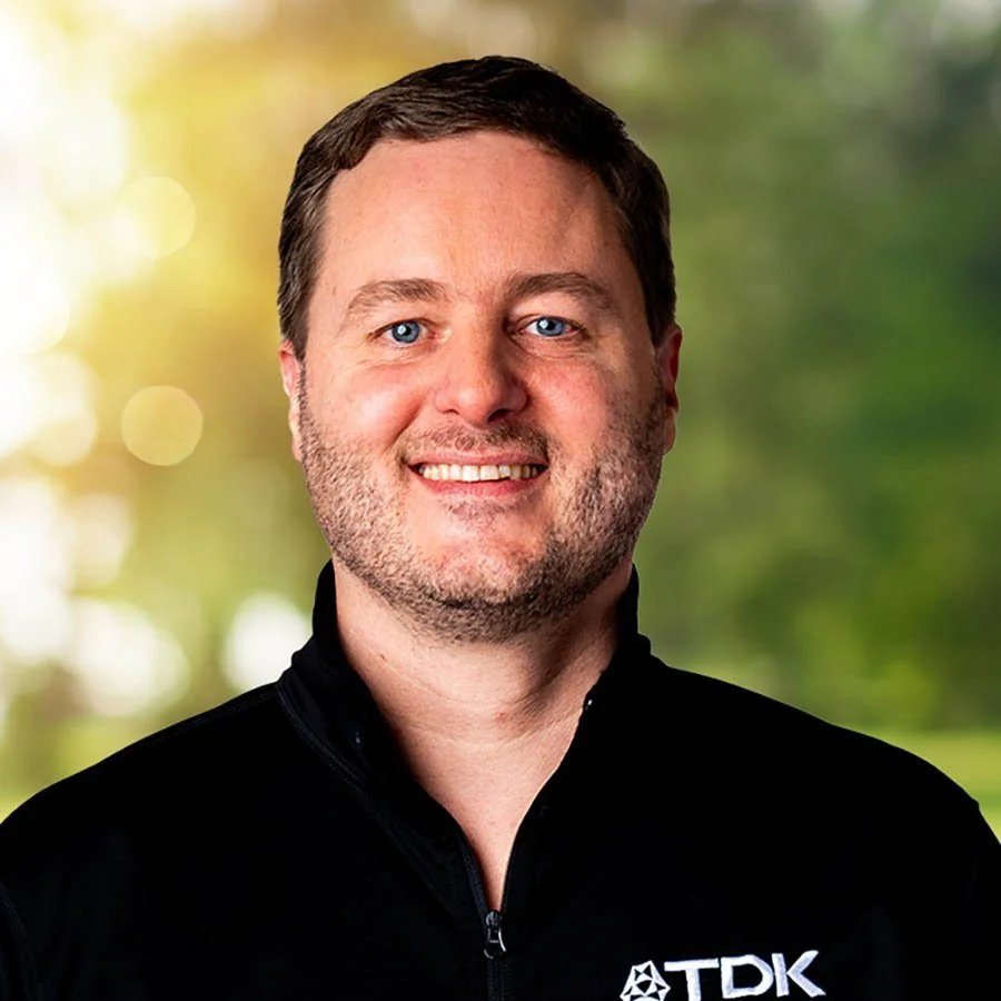A smiling man with short brown hair, blue eyes, and light skin, wearing a black jacket with a TDK logo, outdoors with blurred green trees in the background.