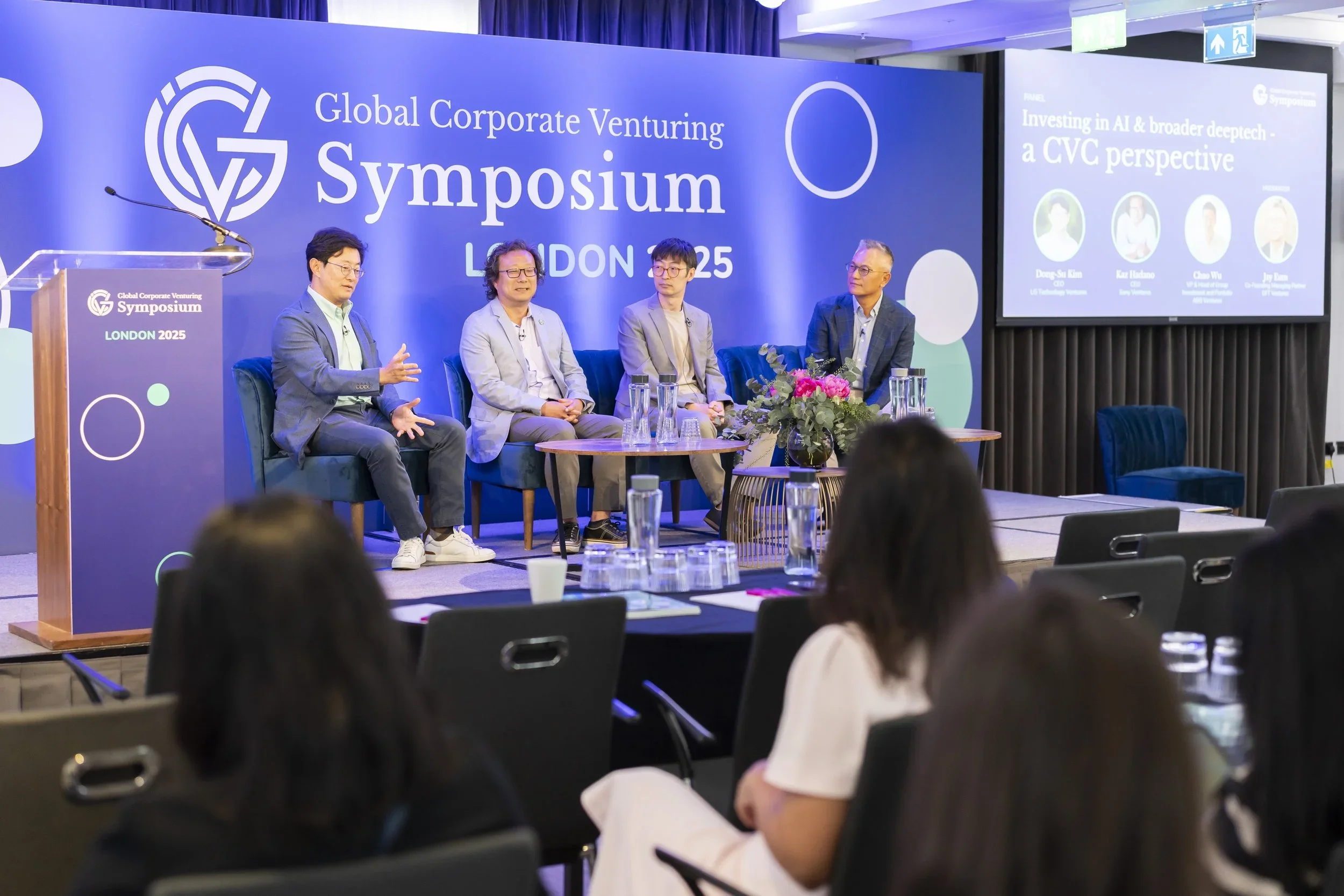 A panel of four men sitting on a stage at the Global Corporate Venturing Symposium in London 2025. The backdrop displays the event name and location. An audience is seated in front, and a screen with a presentation is visible.
