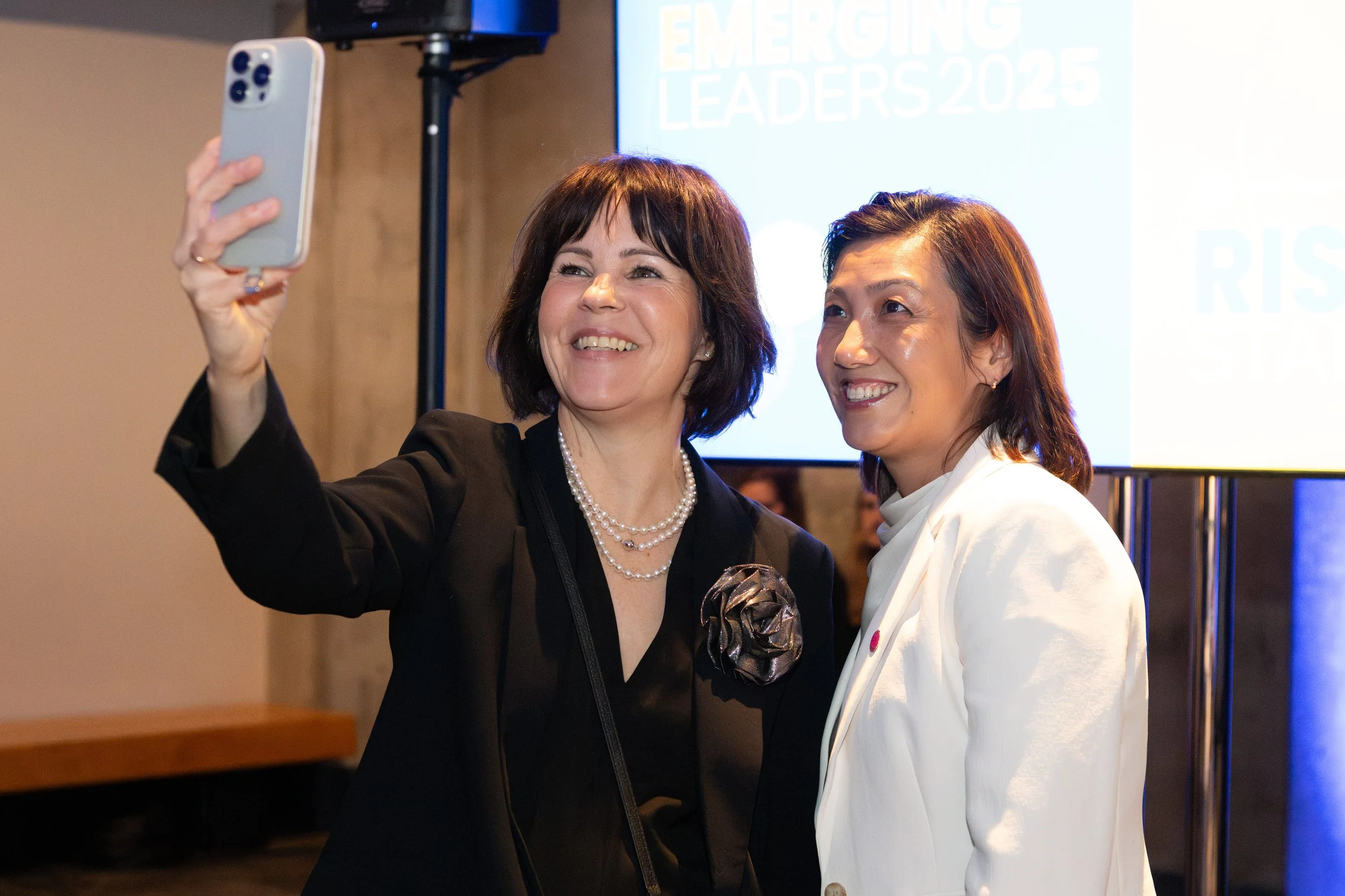 Two women taking a selfie together at an event, with a presentation screen in the background that reads 'Emerging Leaders 2025'.
