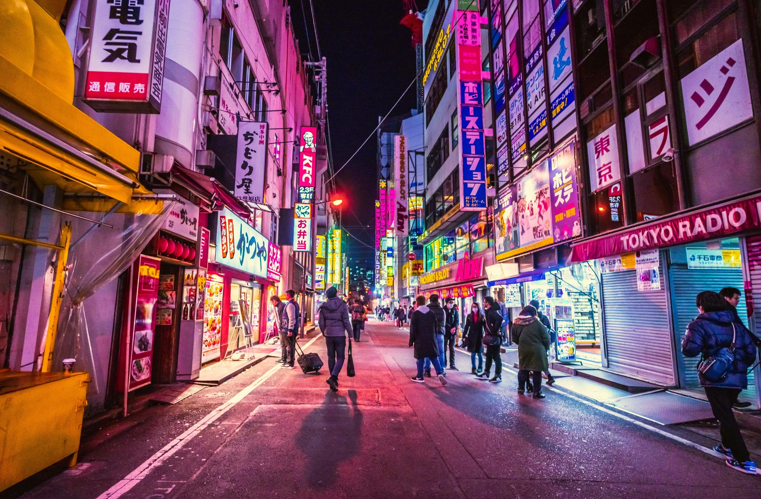 Nighttime street scene in Tokyo with neon signs, shops, and pedestrians.
