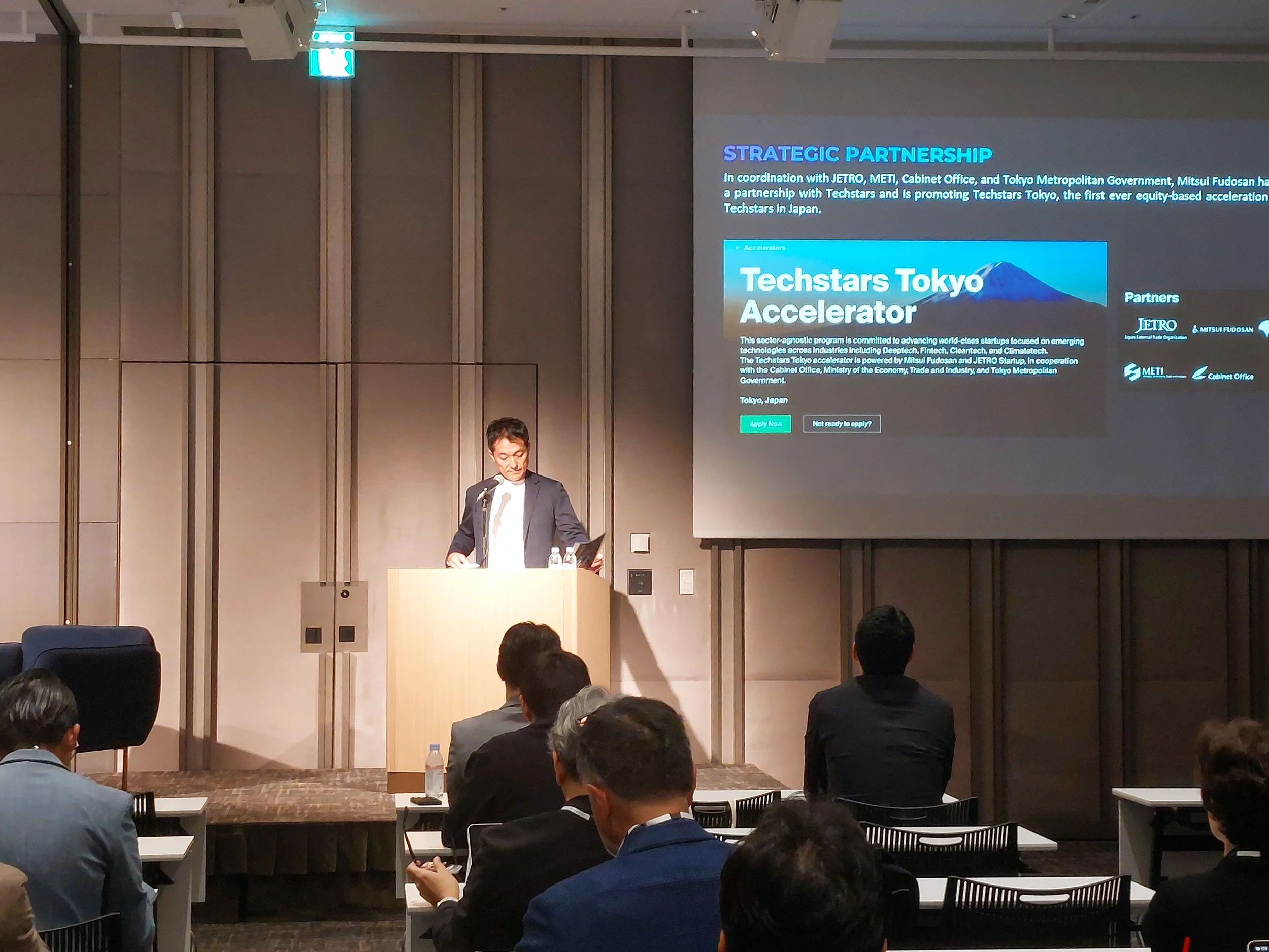 A person standing at a podium giving a presentation in a conference room, with a large screen displaying information about a partnership and accelerator program.