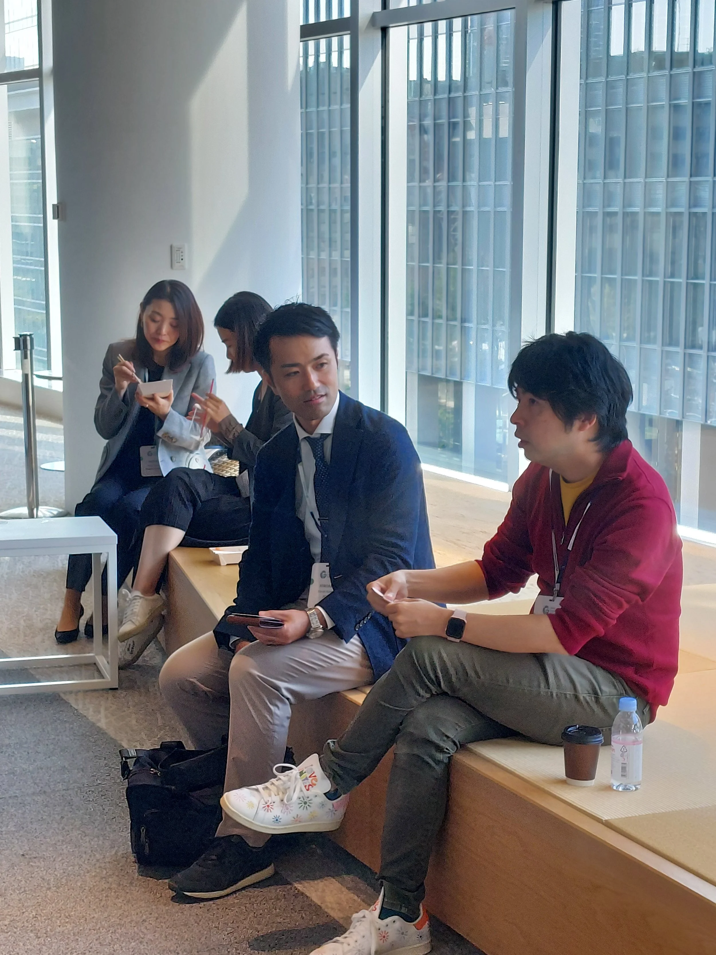 Four individuals sitting in a modern indoor space with large windows. Two women are seated on the left, and two men are seated on the right, engaging in conversation. The women are taking notes, and the men are holding electronic devices. There are water bottles and coffee cups nearby.