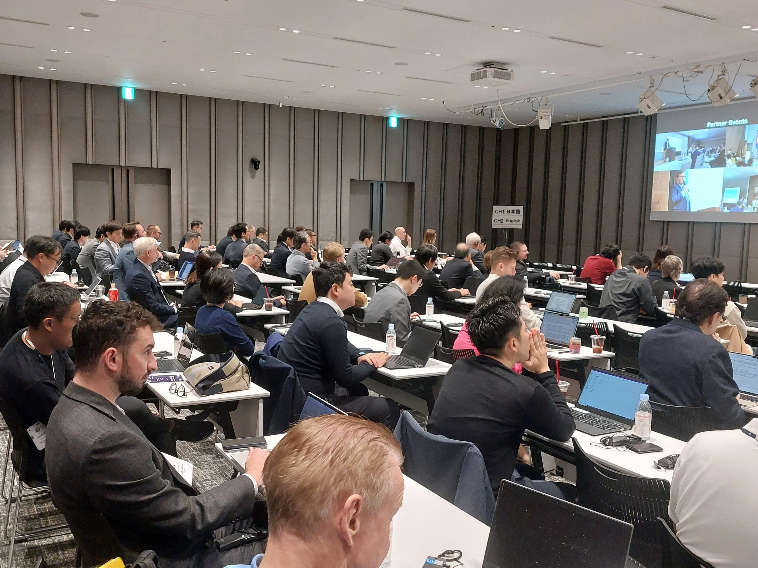 Conference room filled with attending professionals, seated at tables with laptops, listening to a speaker and viewing a presentation on a large screen.