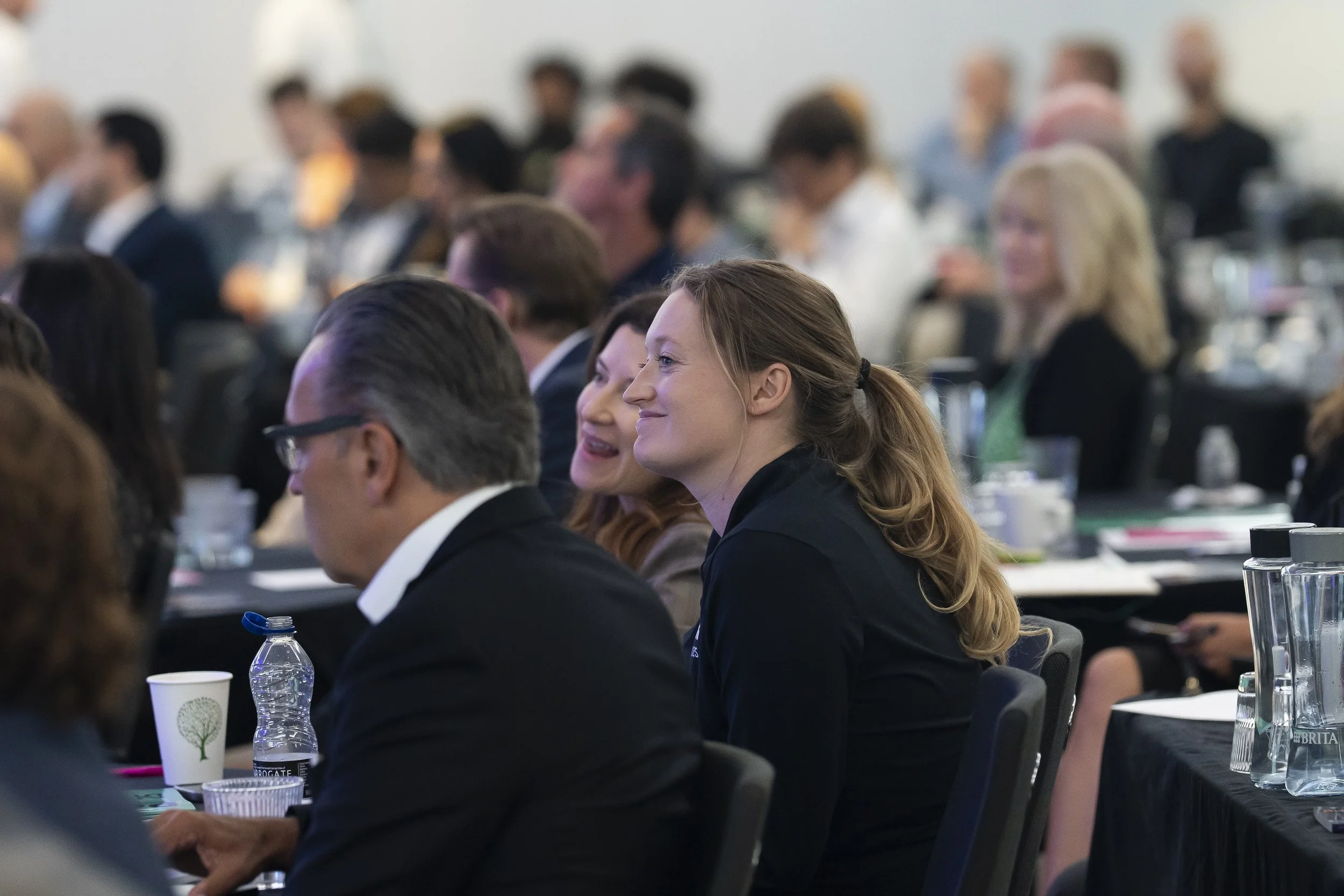 Attendees at a conference sitting at tables, some smiling and listening, with water bottles, paper cups, and notebooks on the tables.