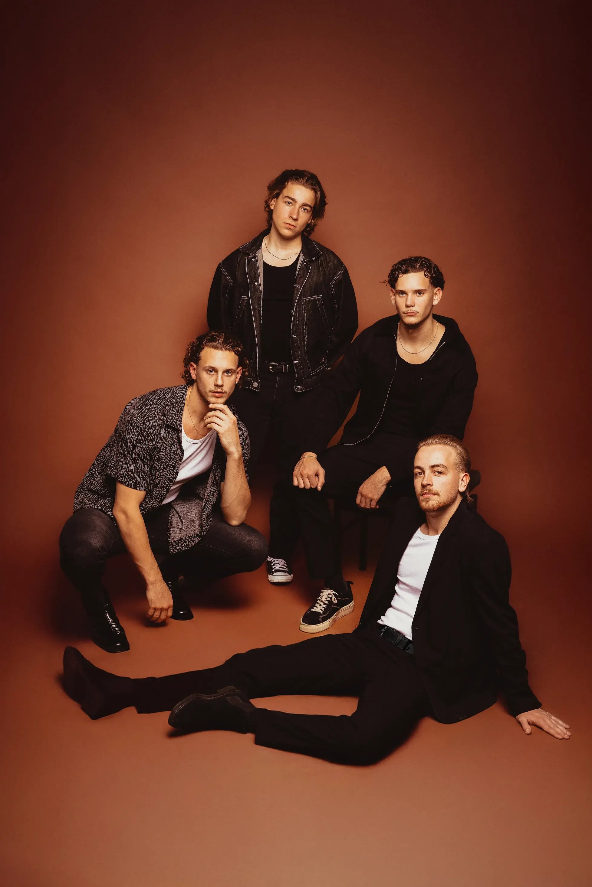 Five young men with styled hair posing against a brown background, wearing casual and stylish clothing, some seated and others standing, in a fashion photoshoot setting.