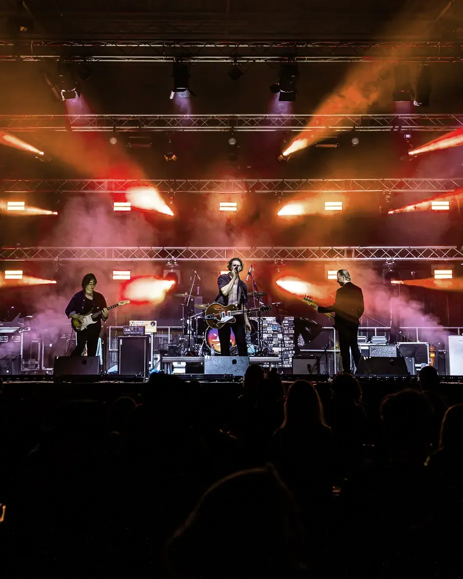 Band performing on stage with bright stage lights and smoke effects, audience in foreground.