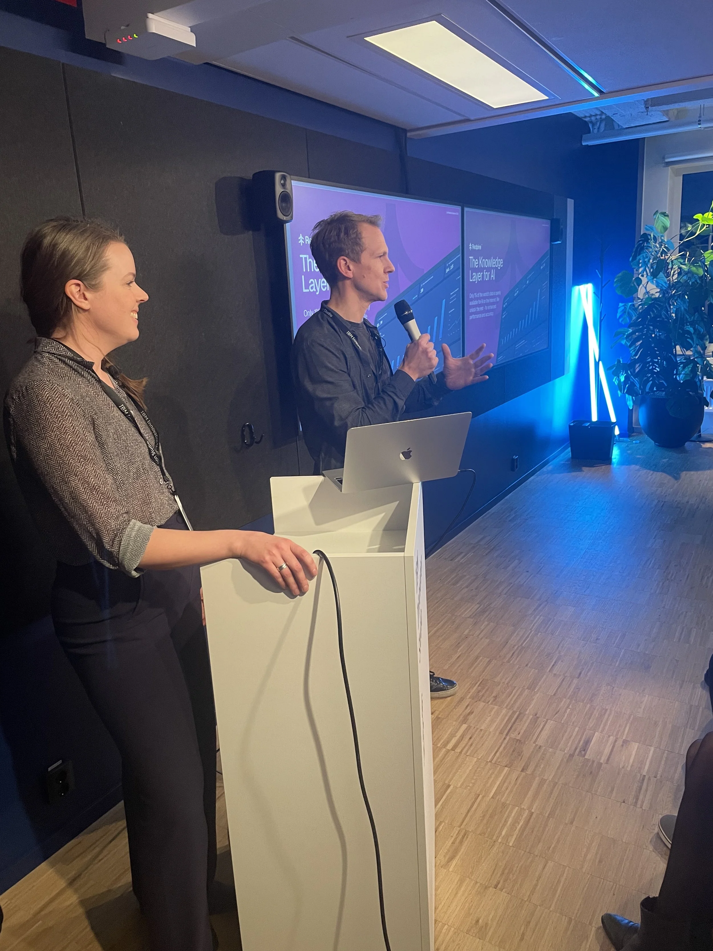 Two people are presenting at an event. One person, a man, is speaking into a microphone with a presentation slide in the background, and a woman is standing beside a podium, listening. The room has a large screen, some blue lighting, and a large potted plant.
