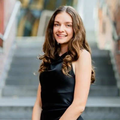 Young woman with long, wavy brown hair smiling outdoors on city stairs, wearing a black sleeveless dress.