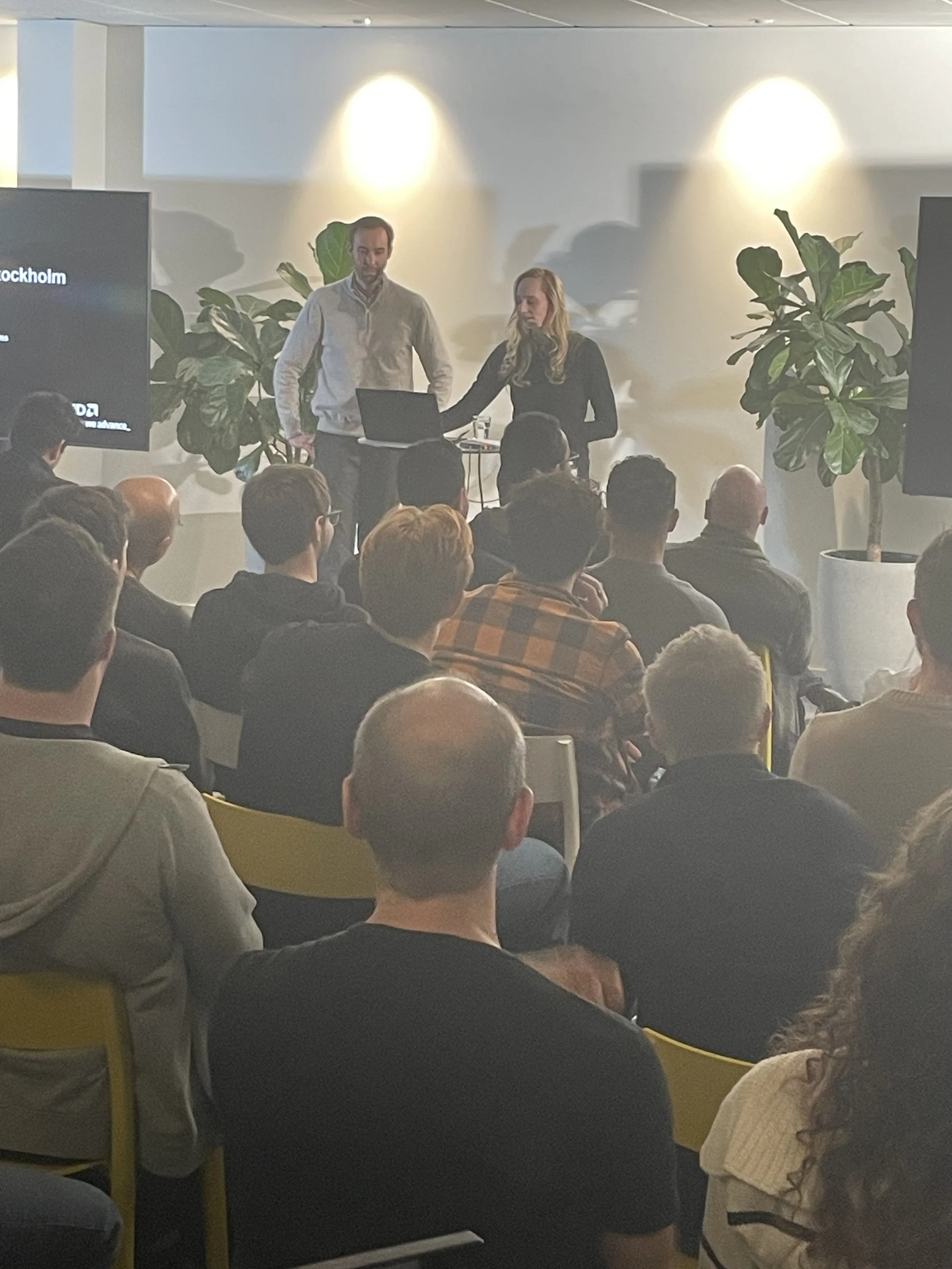 A man and a woman are standing in front of an audience during a presentation or conference, with large plants on either side. The man is wearing a light-colored jacket, and the woman is wearing a black top. They are focused on a laptop on a small table.