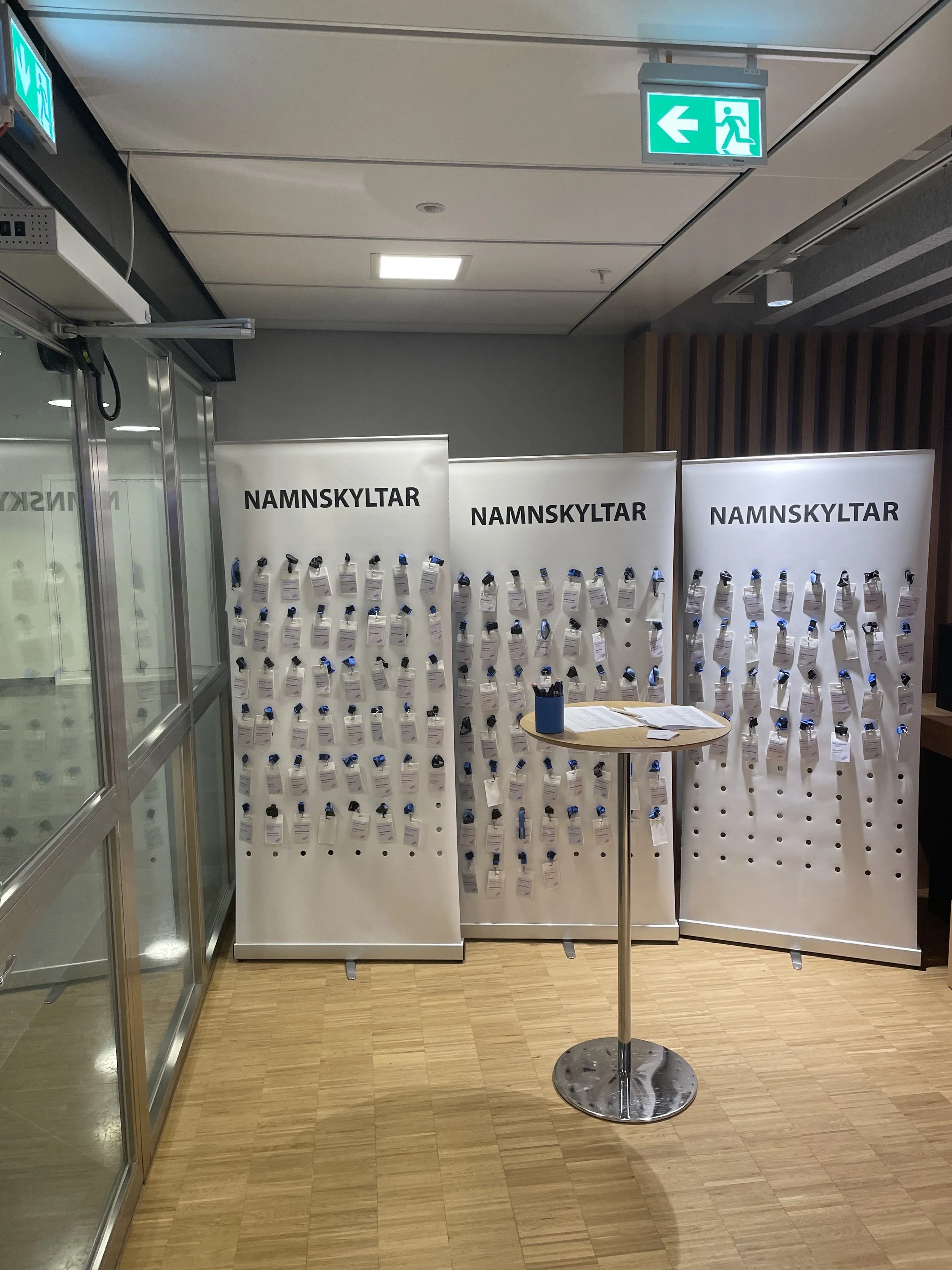 An indoor display with white panels labeled "NAMNSKYLTAR" filled with name tags. A small round table with a blue pen holder and papers is in front of the panels. An emergency exit sign is hanging from the ceiling above, pointing left.