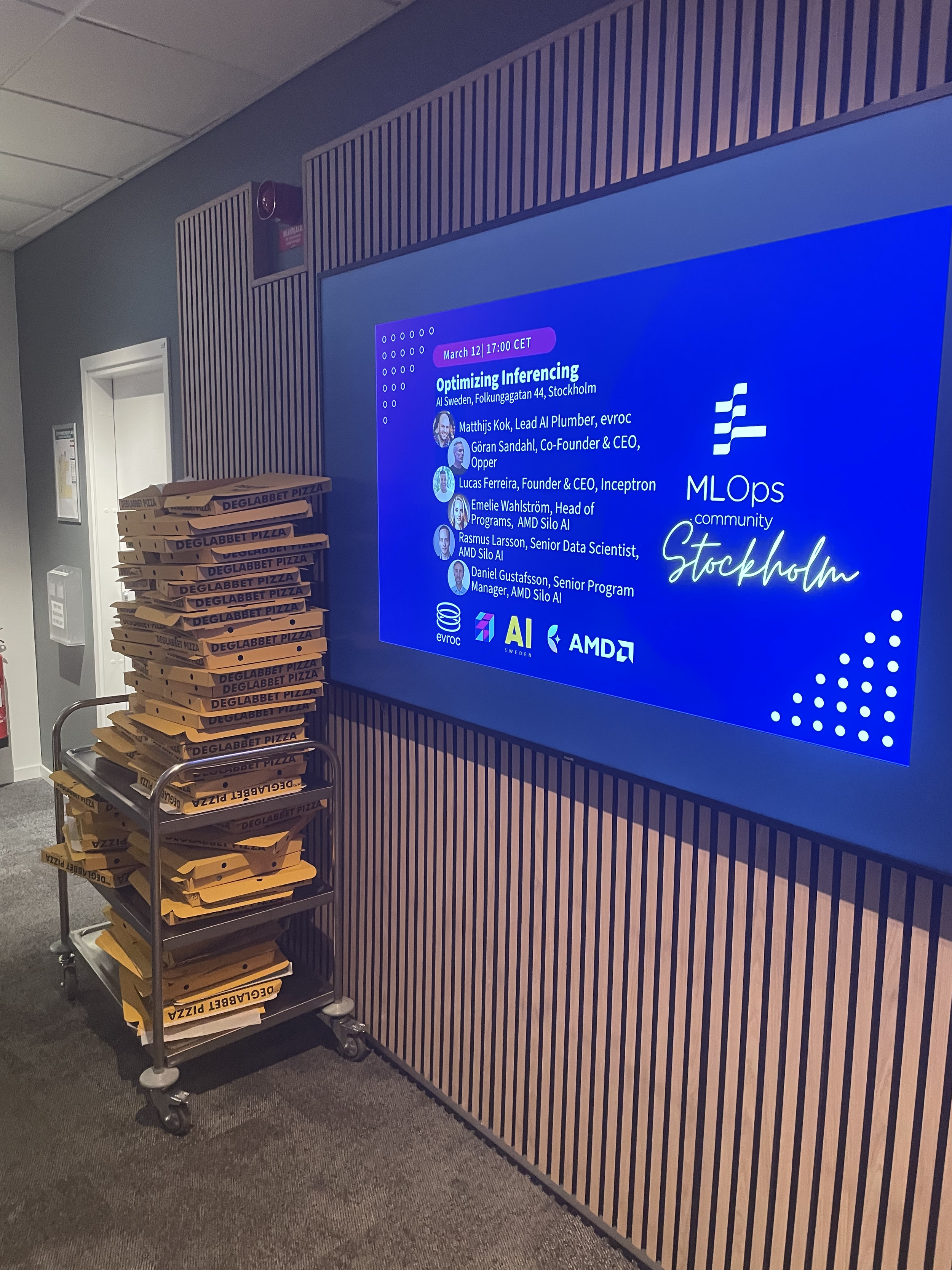 Stack of pizza boxes labeled 'Deglabbat Pizza' on a rolling cart beside a digital conference display with event details and speaker names.