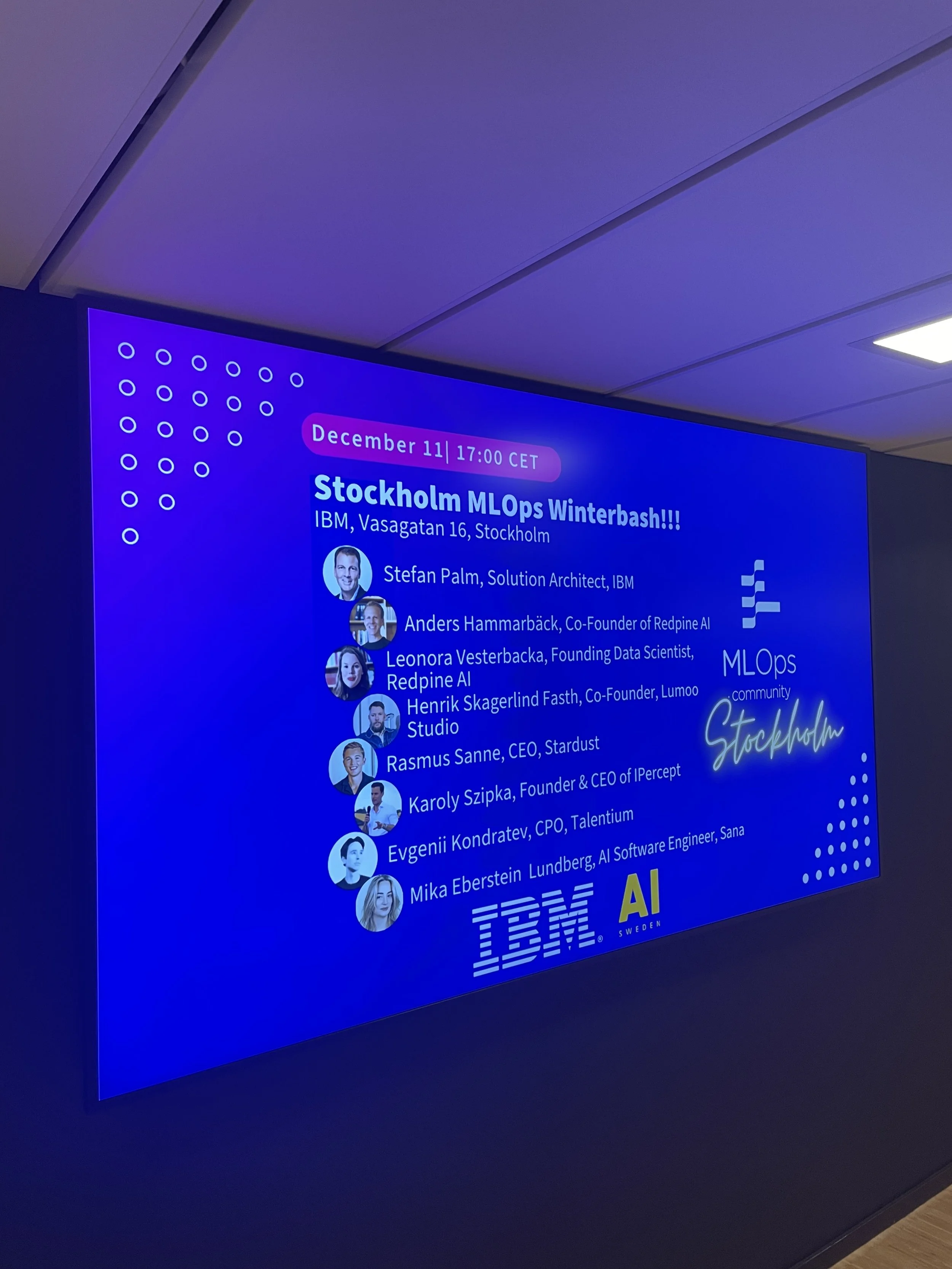 A digital screen displaying information about a tech event called Stockholm MLOps Winterbash. It shows the date and time, December 11 at 17:00 CET, with details of speakers from IBM including Stefan Palm, Anders Hammarbäck, Leonora Vesterbacka, Henrik Skagerlind Faste, Rasmus Sanne, Karoly Szlpa, Evgenii Kondratiev, and Mika Eberstein. The screen has a blue background with white text, images of speakers, logos for IBM and AI Sweden.