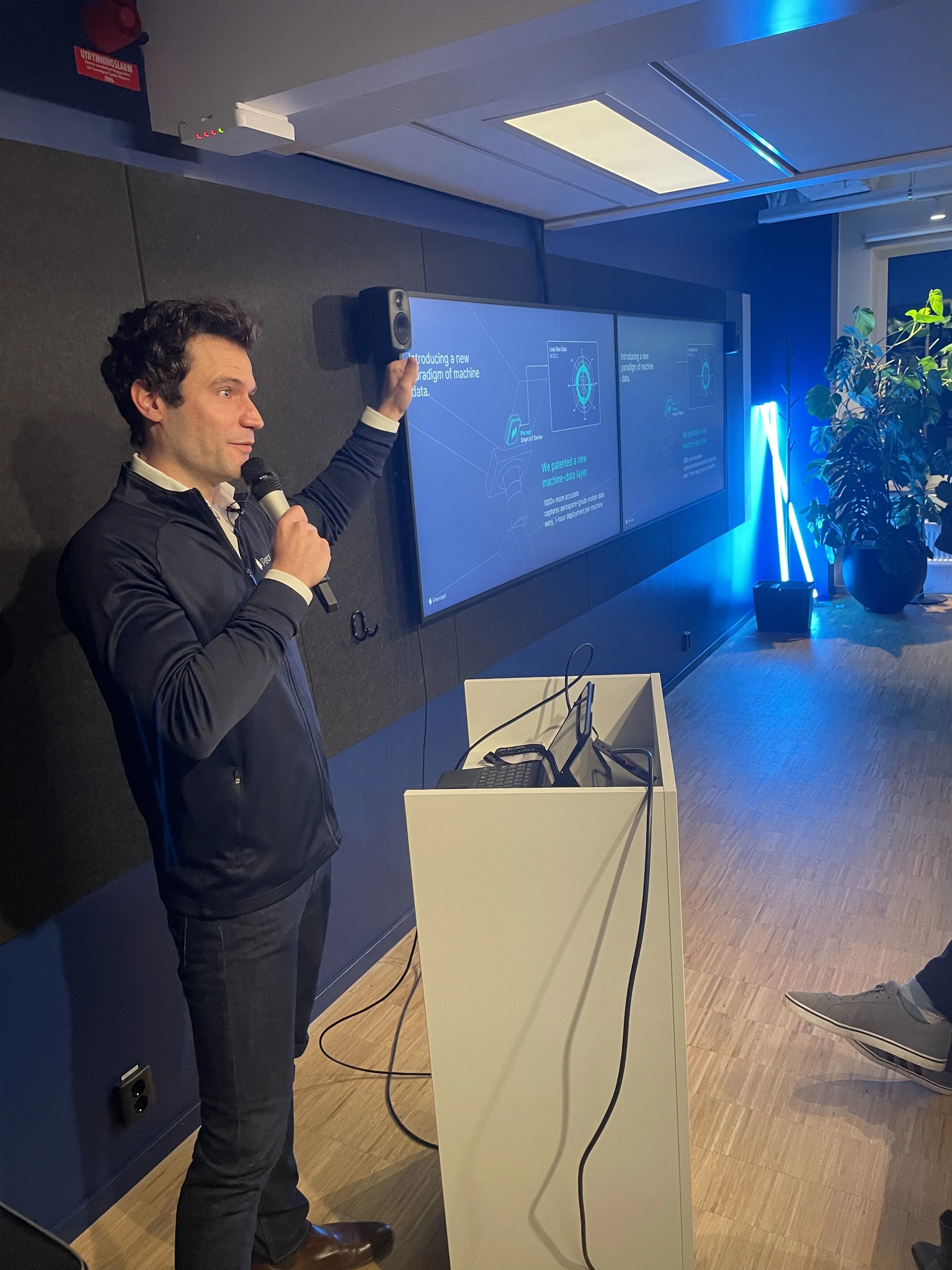 A man holding a microphone and gesturing with his hand, standing in front of a presentation screen in a conference room with blue lighting and plants.