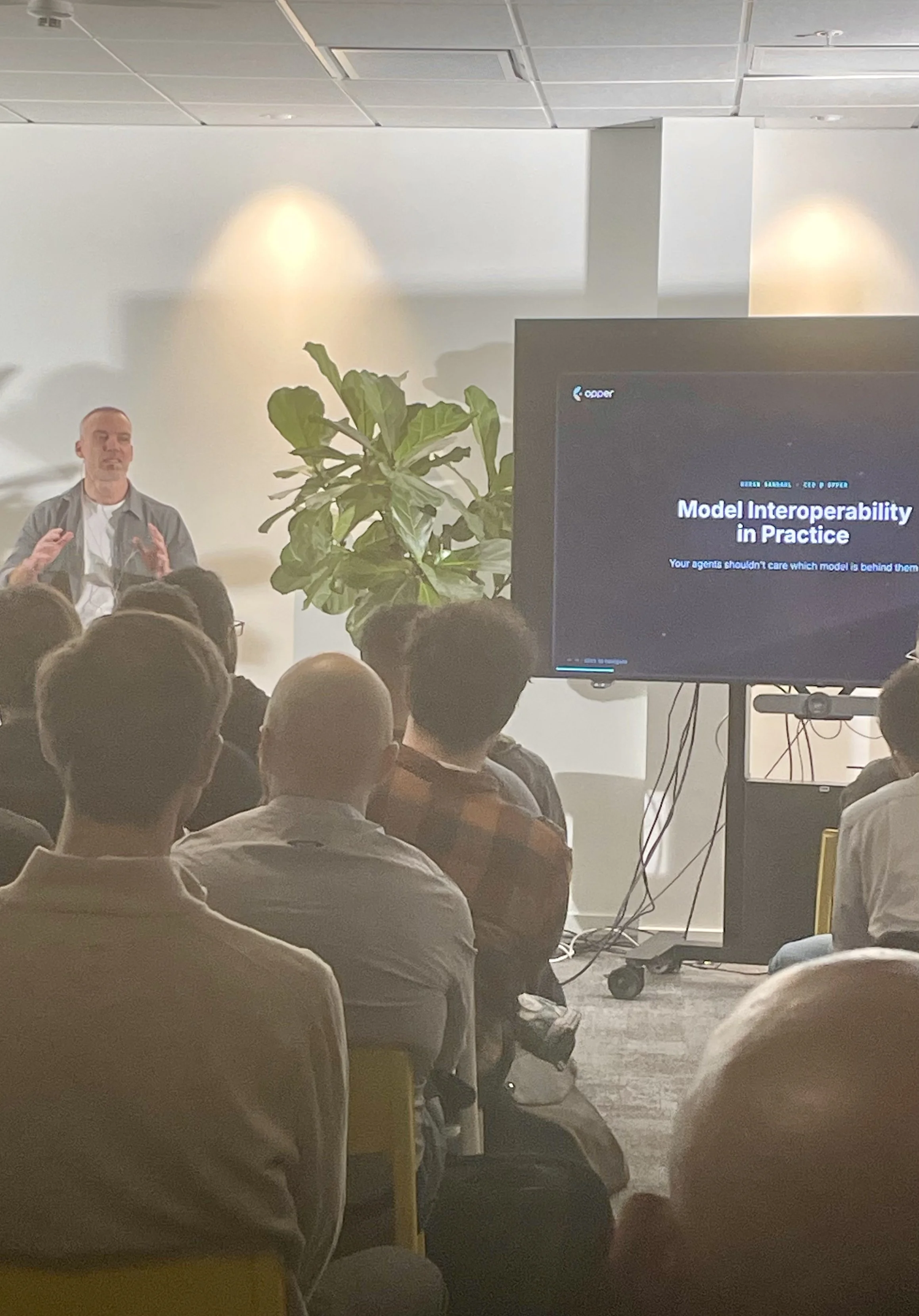 A man is giving a presentation in a conference room. The presentation screen reads 'Model Interoperability in Practice' with a subtitle 'Your agents shouldn't care which model is behind them.' Audience members are seated, facing the speaker and screen, with a large plant positioned near the front of the room.