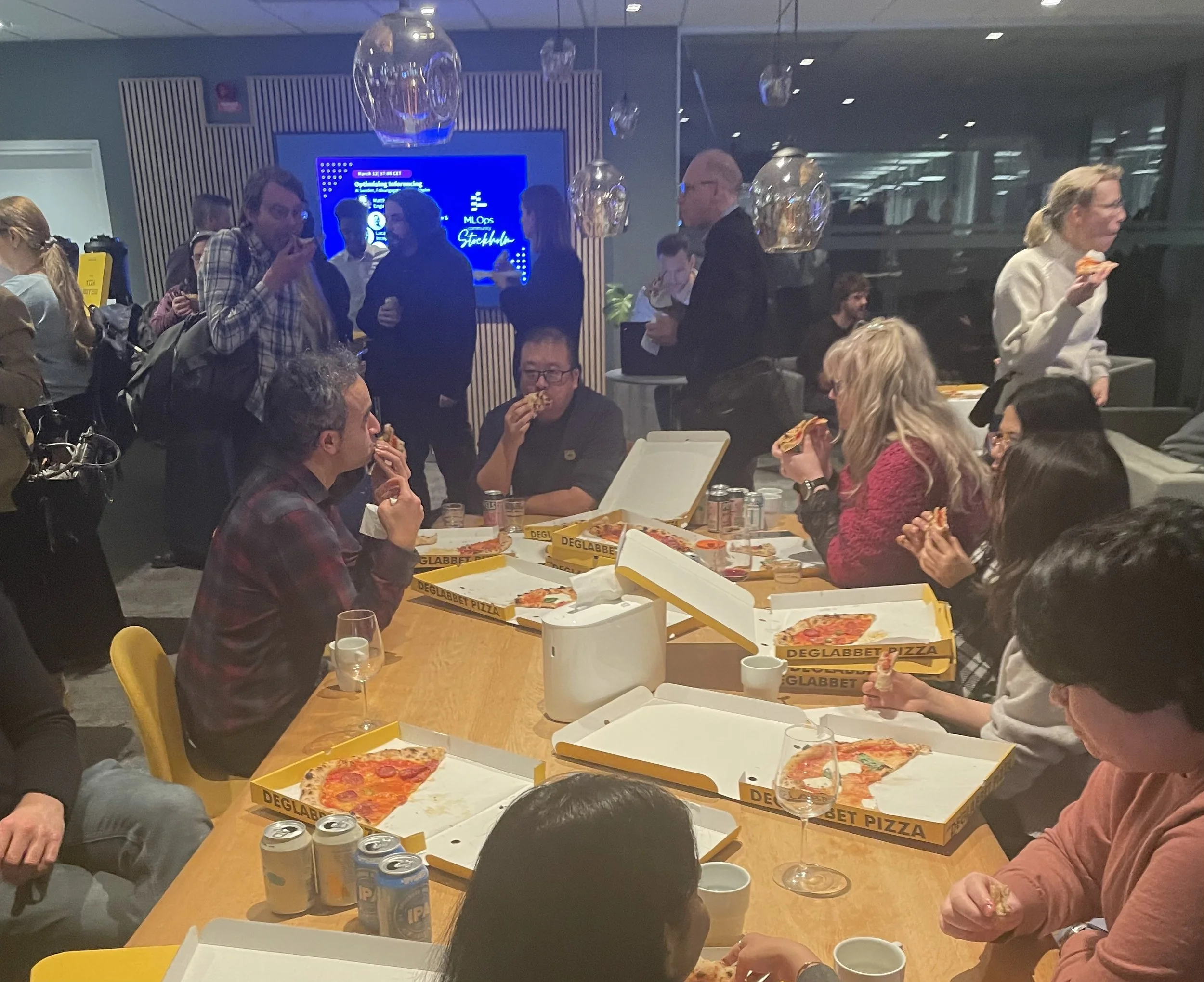 A group of people gathered in a room eating pizza and drinking from cans and wine glasses, with a large blue digital screen in the background displaying event information.