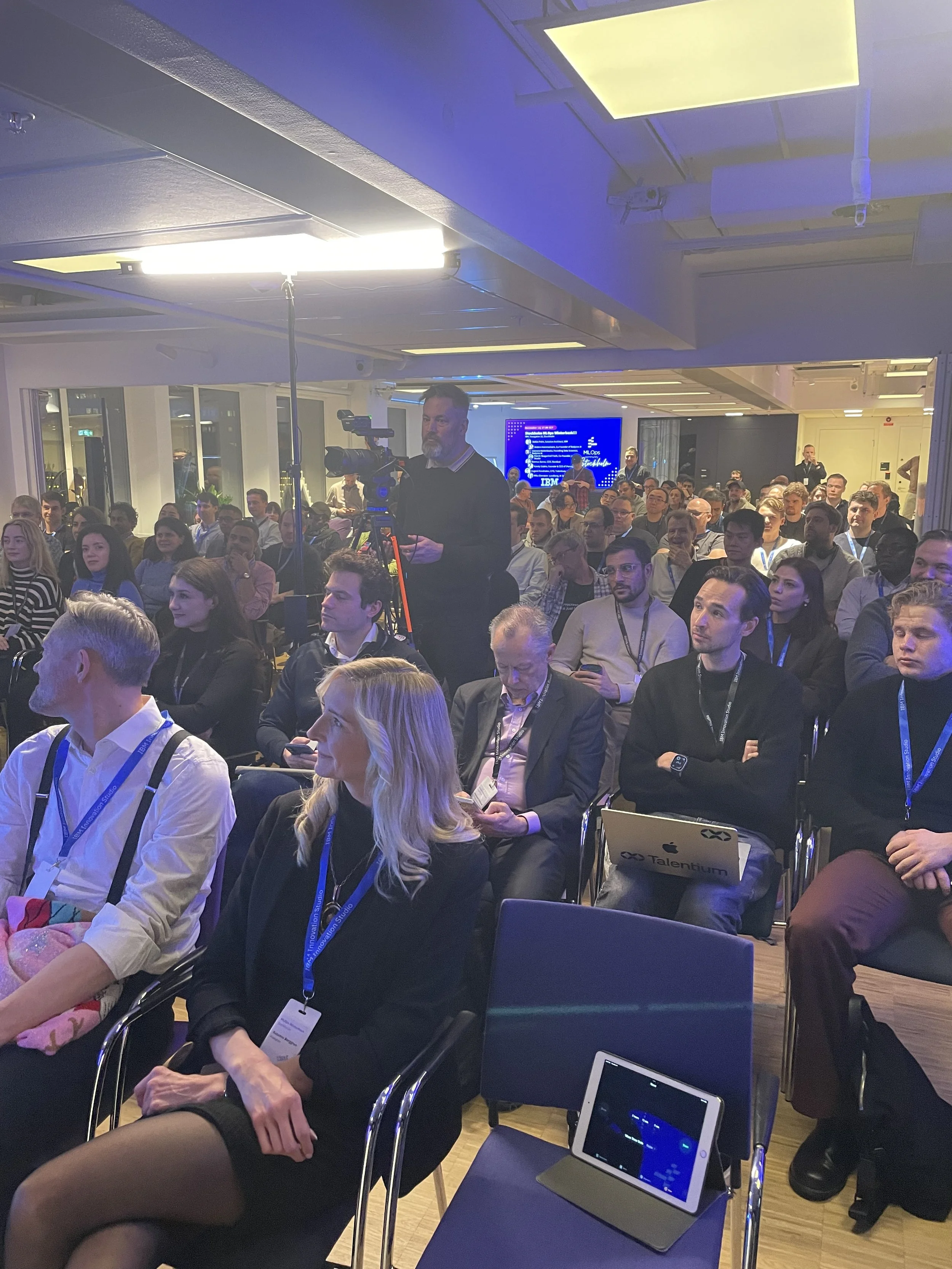 A large group of people attending a conference or seminar, seated in rows and facing forward. Some attendees are using electronic devices, and a camera operator is capturing the event. There is a large screen displaying information in the background.