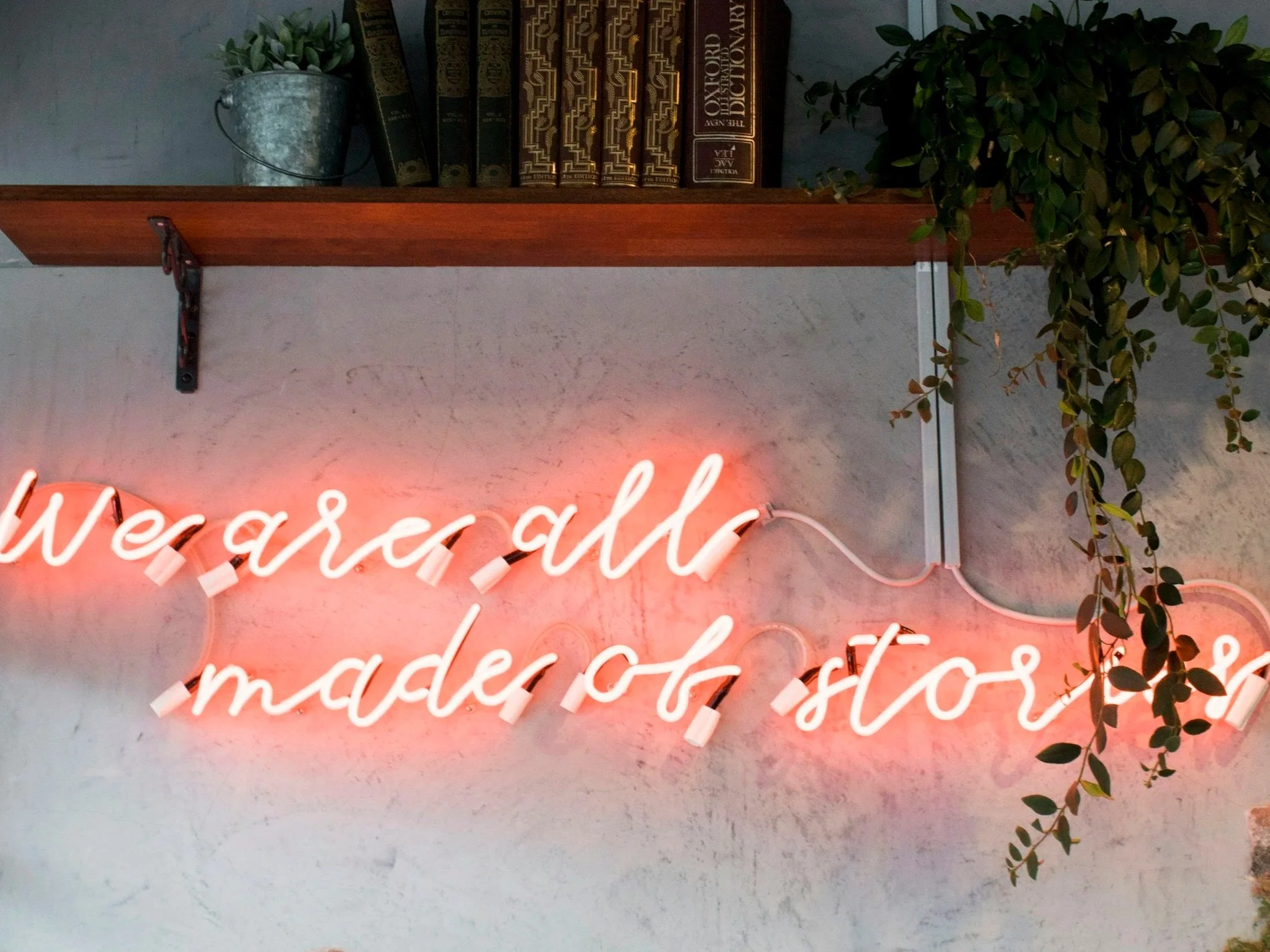Neon sign on wall reading 'wear all made of stars', with a wooden shelf holding books, a small metal bucket with a plant, and hanging greenery.