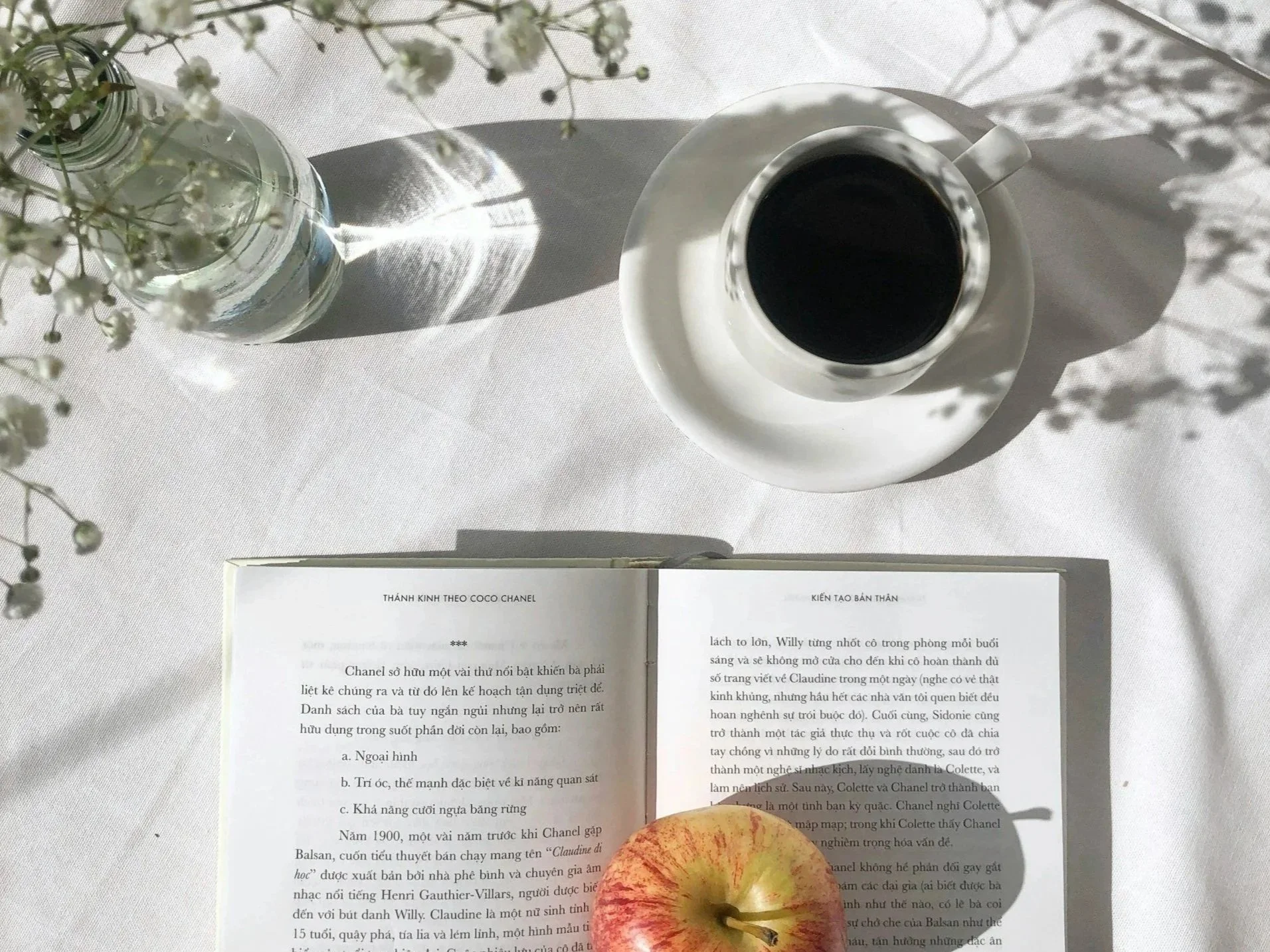 A top-down view of a white table setting with a vase of white flowers, a cup of black coffee on a saucer, an open book with Vietnamese text, an apple, and shadows cast by the flowers and objects.