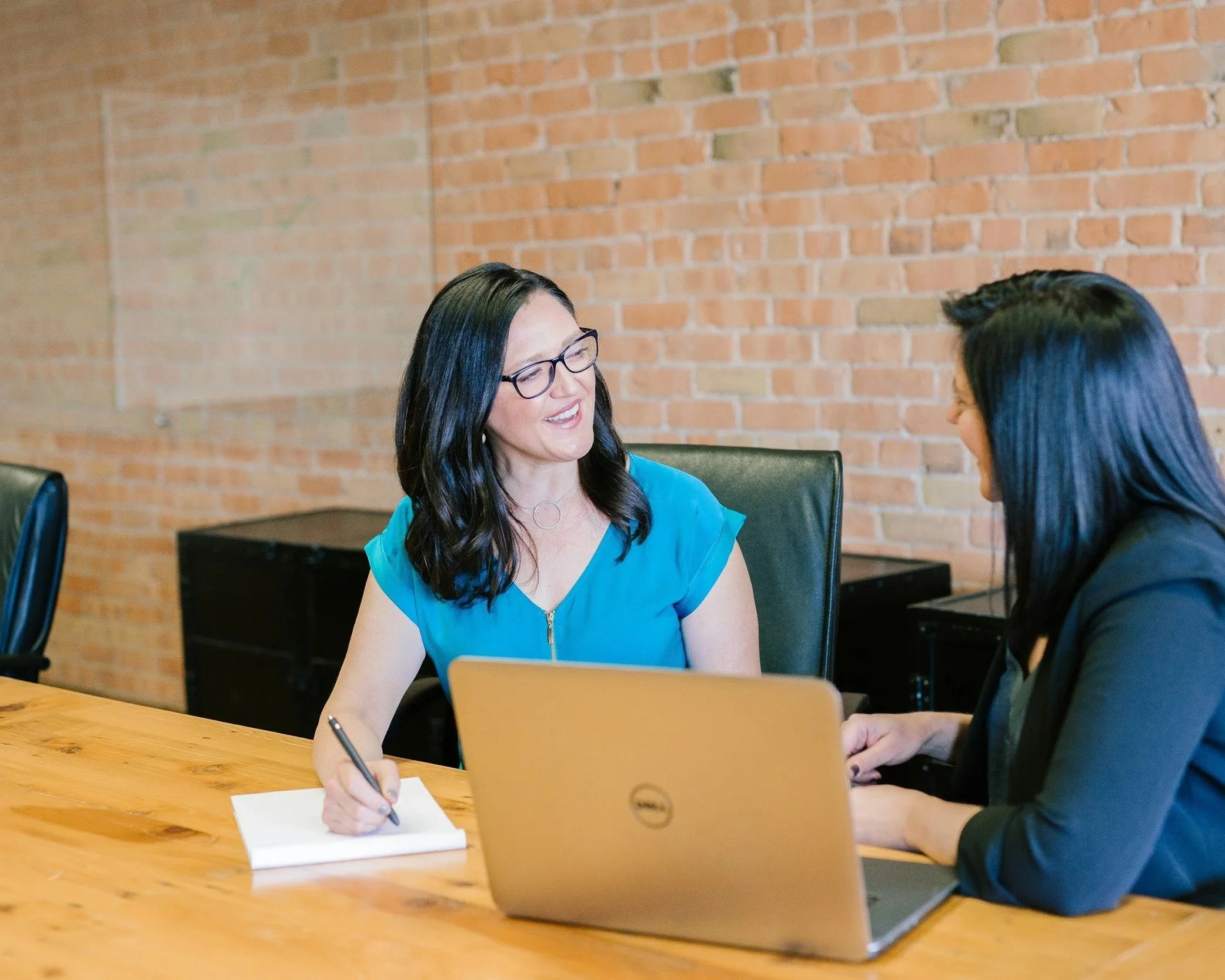 Two women sitting at a wooden table having a conversation in a modern office space with exposed brick walls. One woman is taking notes with a pen, while the other has a laptop open in front of her.