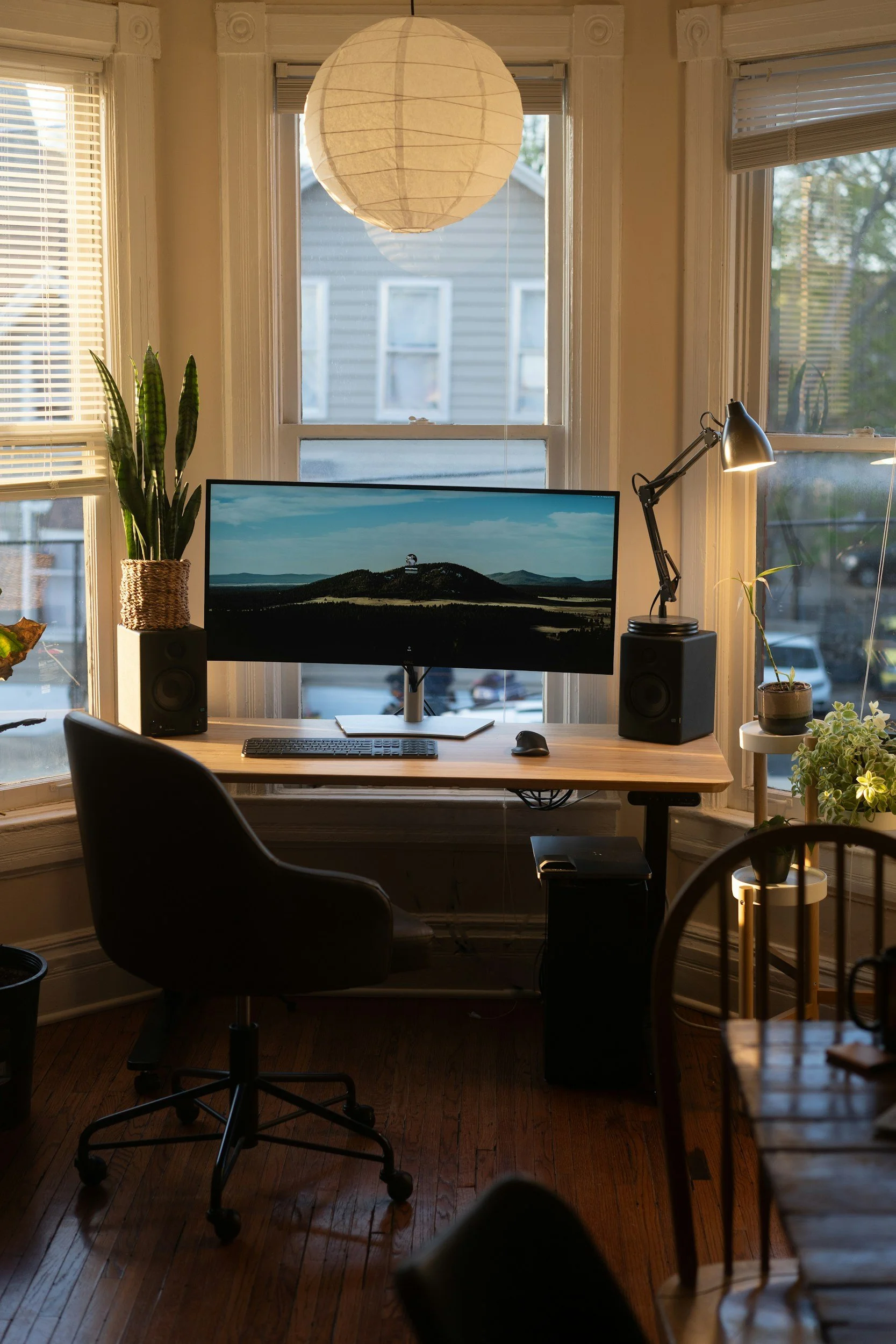 Home office with a desk in front of a window, featuring a computer monitor, a keyboard, a mouse, a desk lamp, a potted plant, and speakers, illuminated by natural light and a ceiling paper lantern.