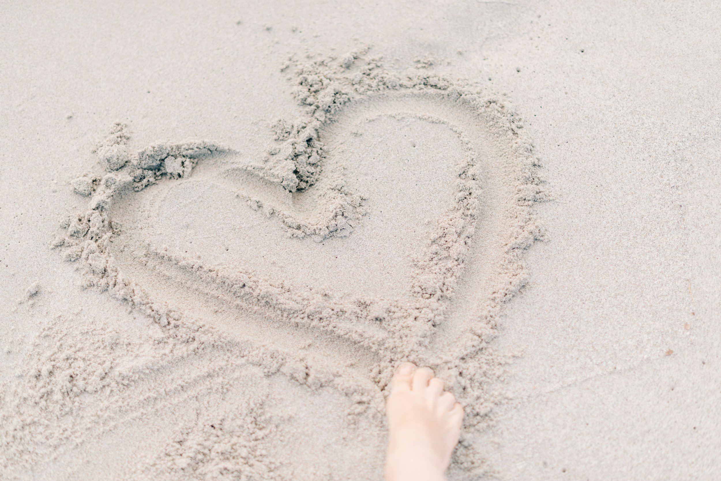 A person's foot is walking on the sand toward a heart shape drawn in the sand, with a few footprints around.