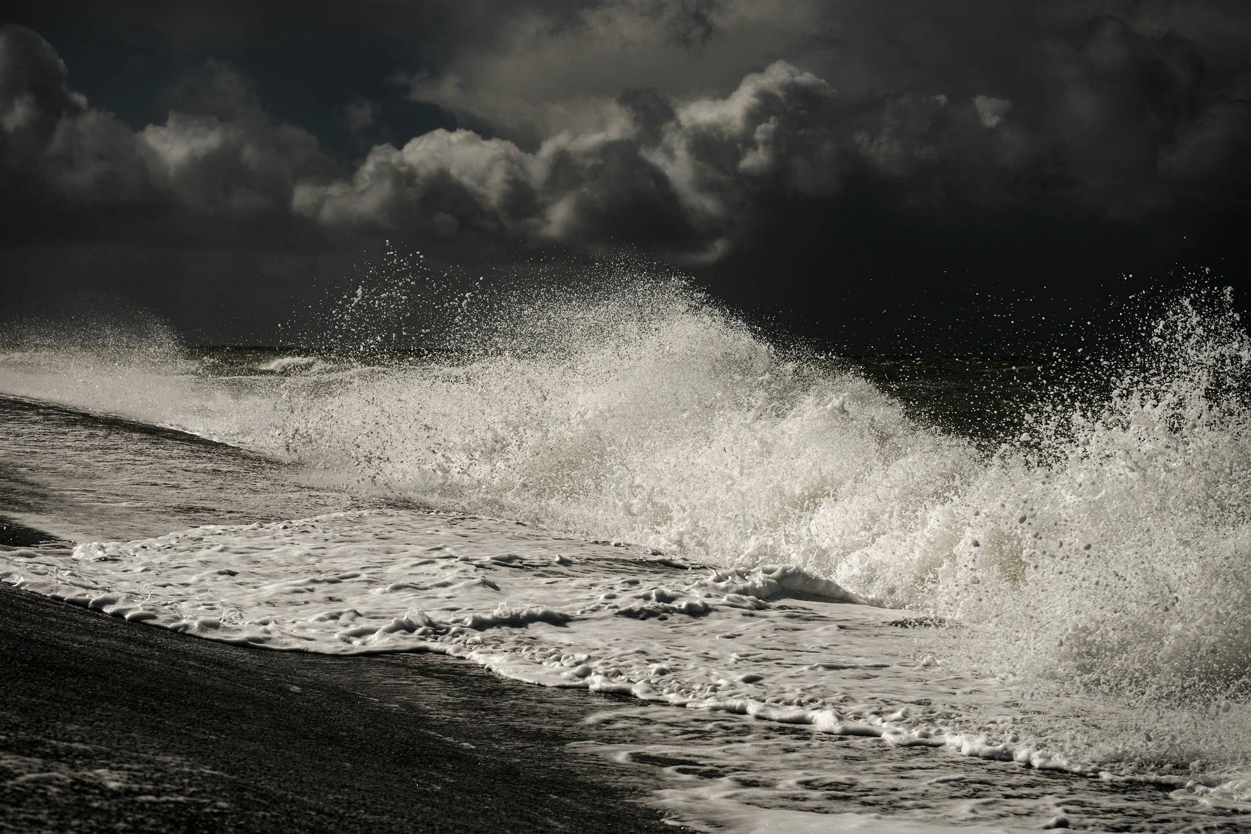 Dark stormy sky over ocean waves crashing on a black sandy beach.