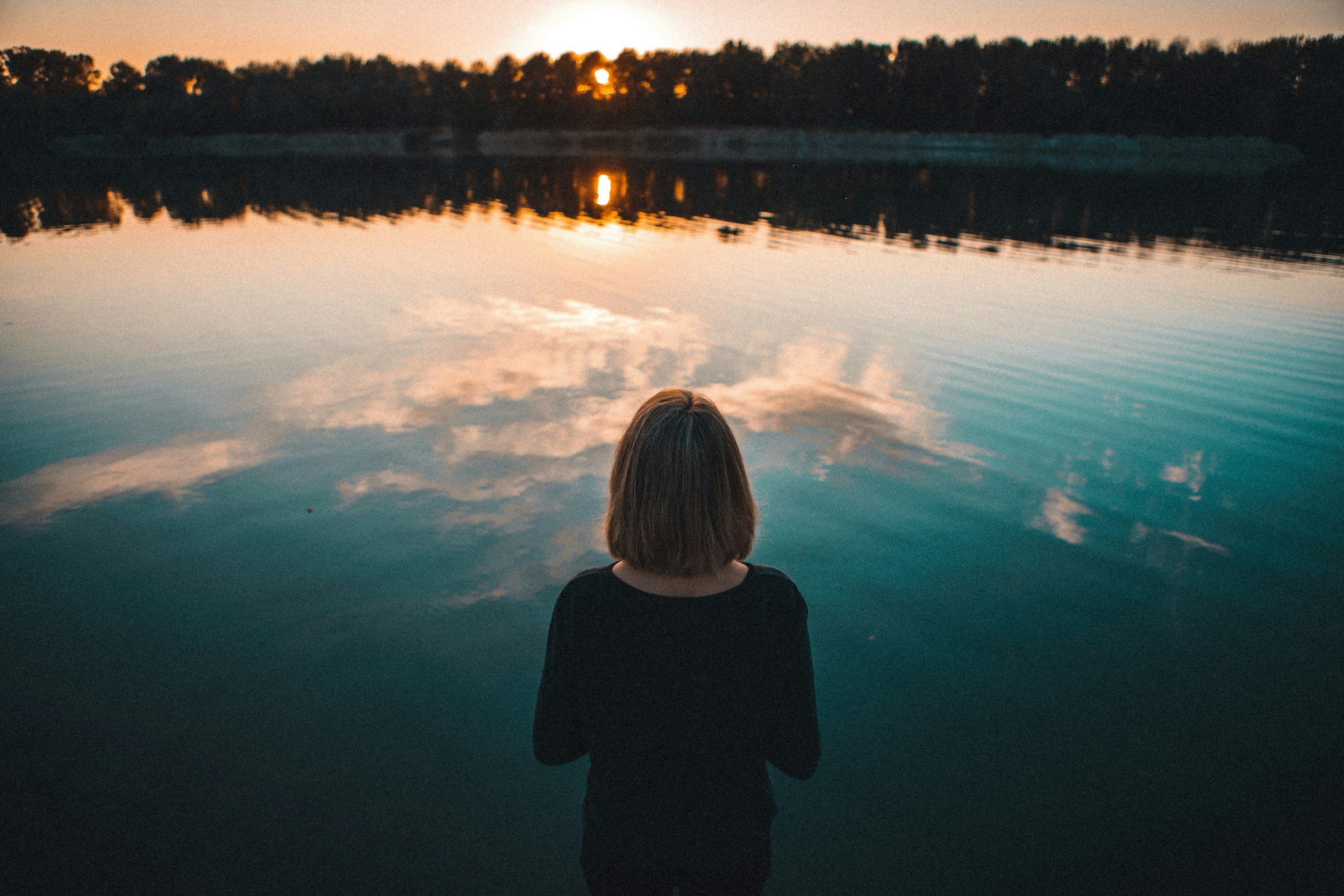 A person with shoulder-length hair standing by a calm body of water at sunset. The sky and water reflect the warm colors of the setting sun, with trees in the background on the horizon.