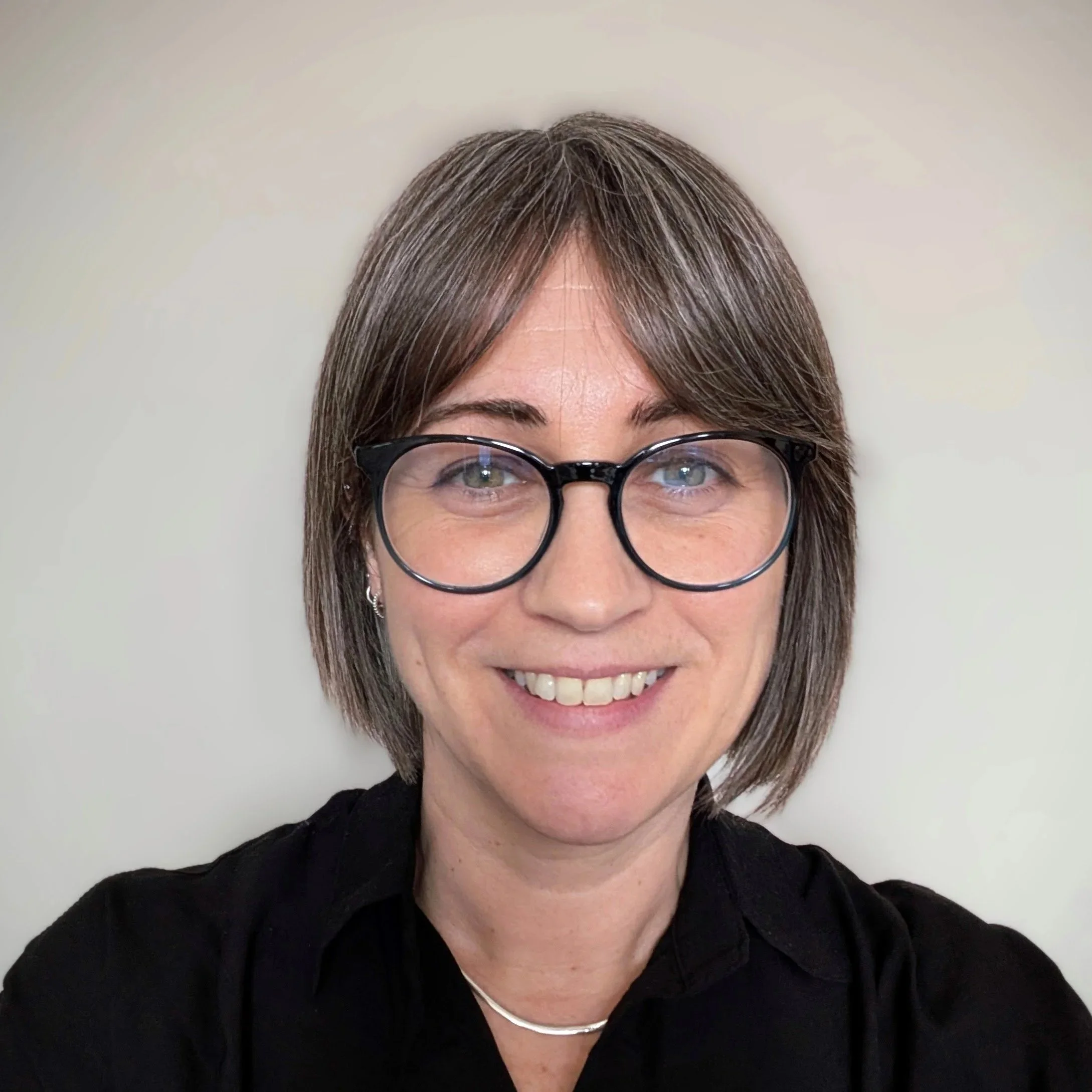 Close-up of a woman wearing glasses, with short brown hair, smiling, against a plain light-colored background, wearing a black top and a necklace.