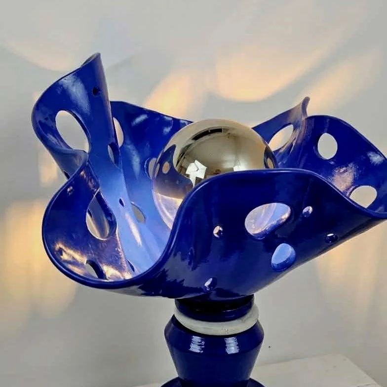 Sculpture lumineuse en argile bleu avec une ampoule à calotte dorée à l'intérieur, placée sur une table blanche.