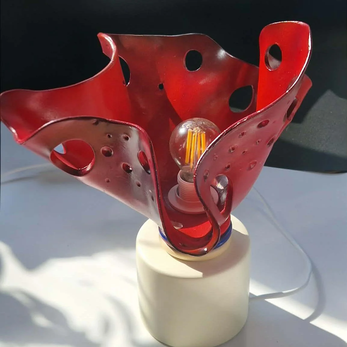Petite lampe avec un abat-jour rouge en argile perforé et une ampoule suspendue au centre, montée sur une base blanche.