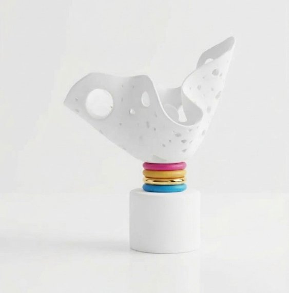 Une sculpture abstraite en forme d'élément blanc avec des perforations, posée sur un support blanc, et décorée de petits anneaux colorés en haut.