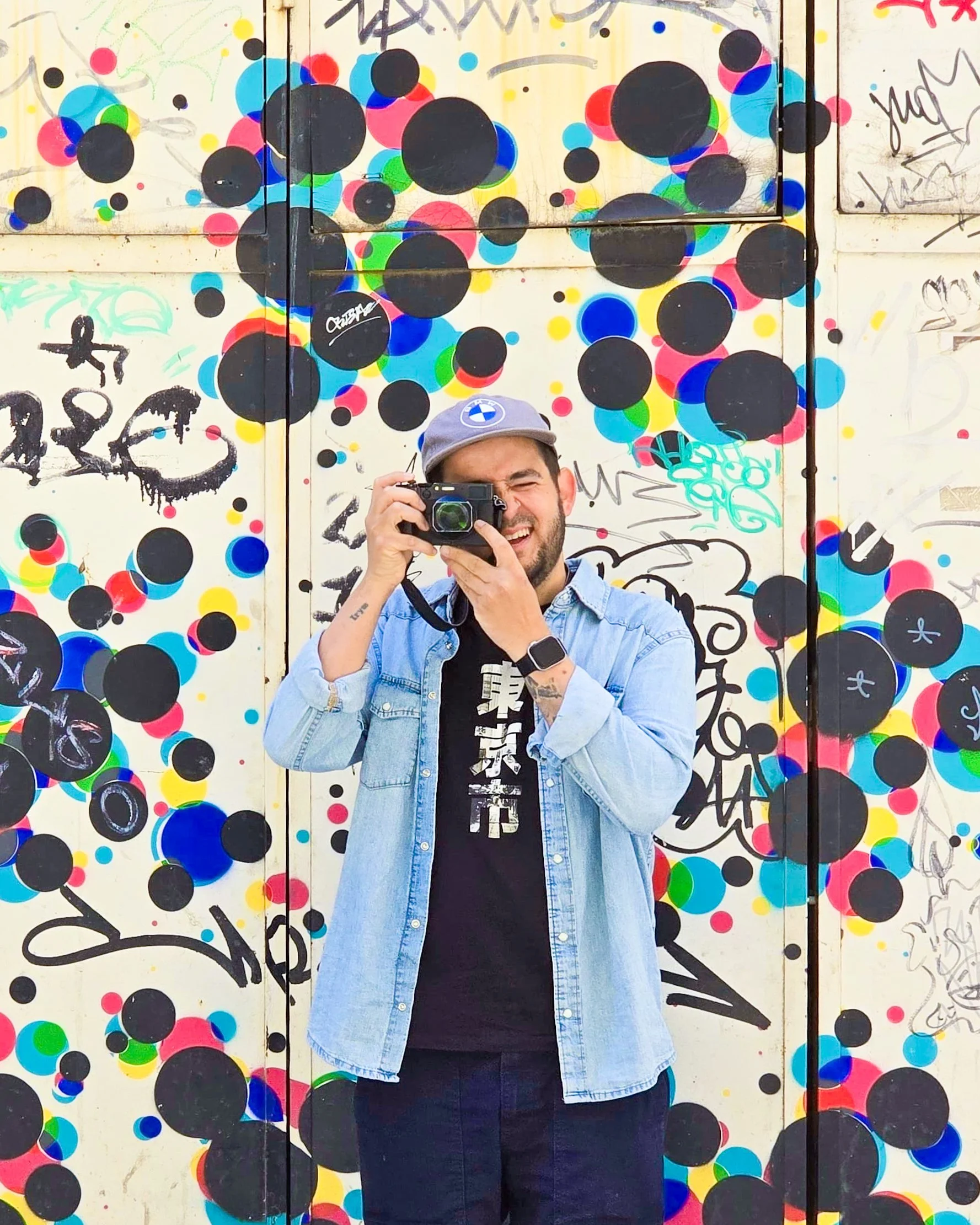 A man with a gray cap and a black shirt is taking a photo of himself with a camera. The background is a colorful graffiti mural with various shapes, black dots, and abstract scribbles.