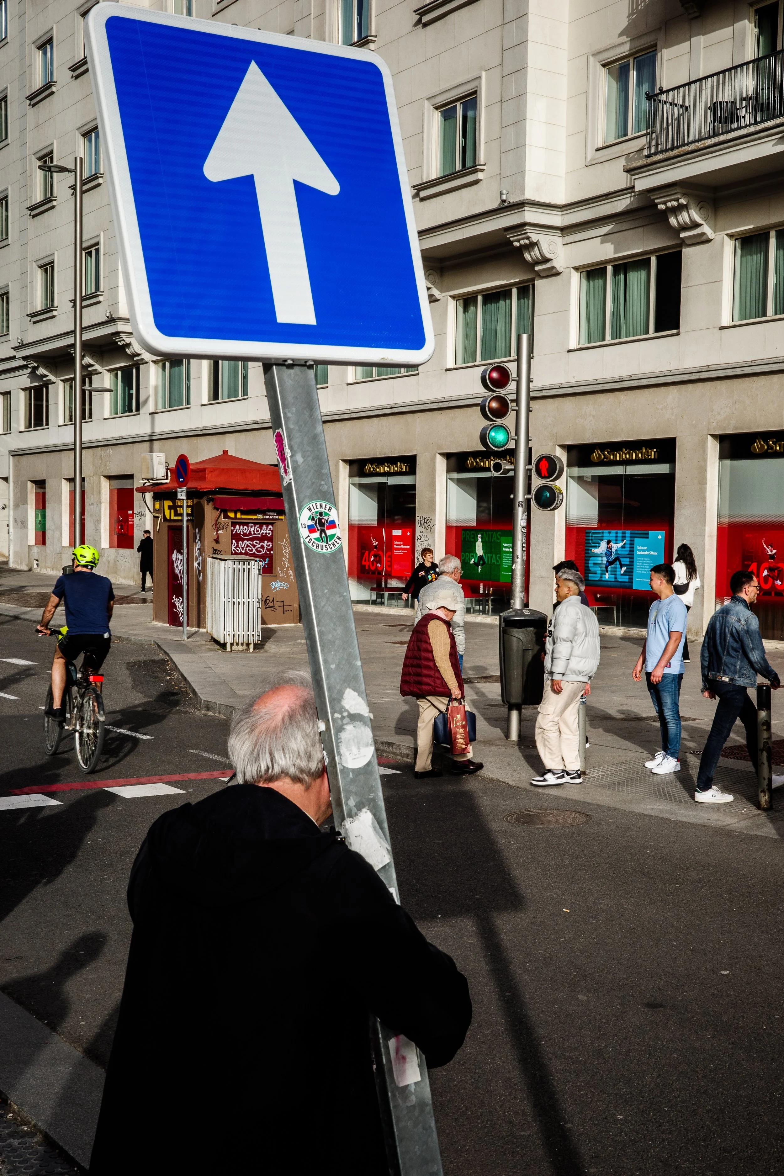 Street scene with a blue upward arrow traffic sign in the foreground, pedestrians crossing the street, cyclist riding on the street, building with shops and apartments in the background, and a traffic light showing green.