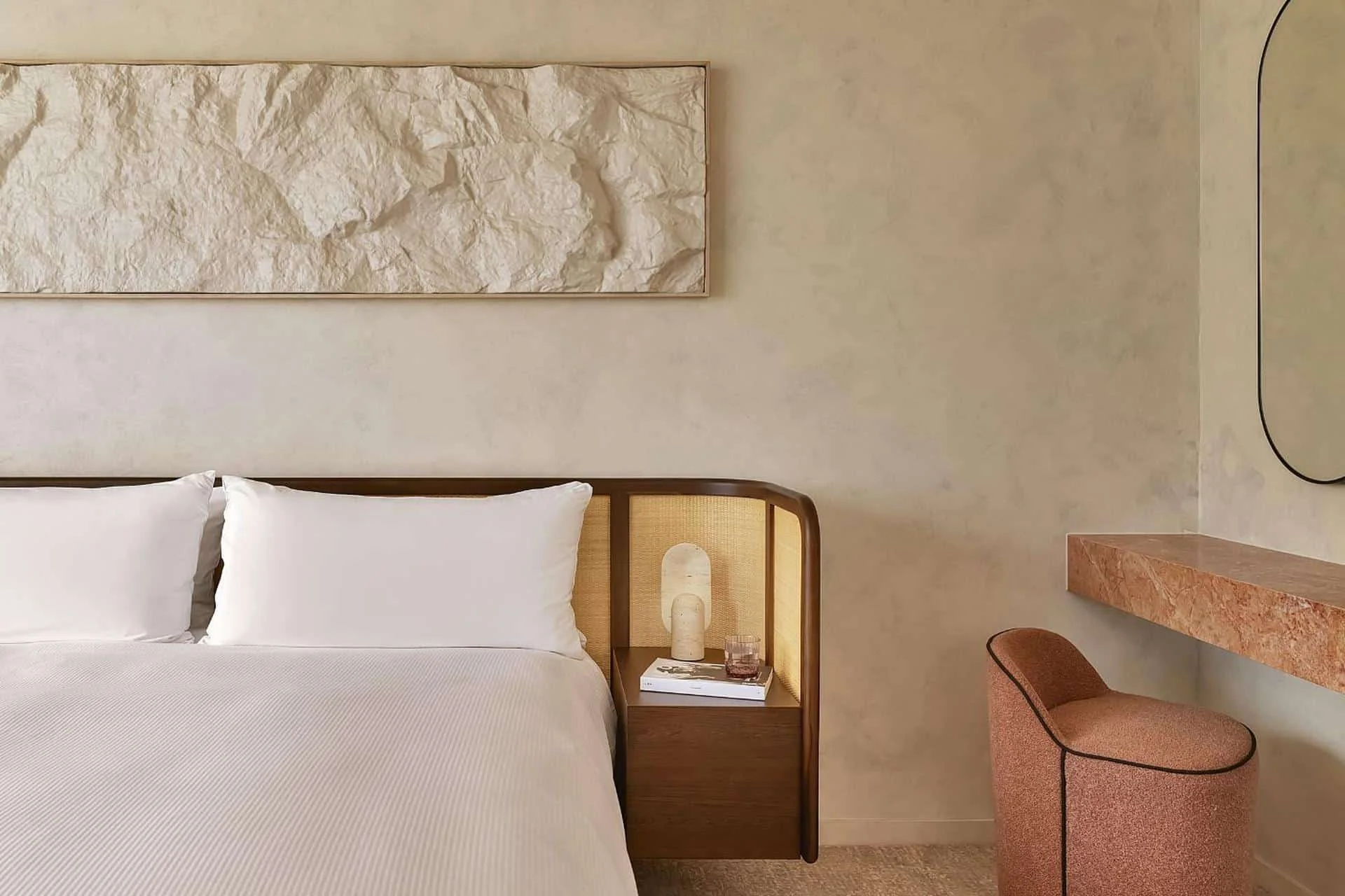 A bedroom with a bed, bedside table with a lamp, books, and a glass of water, a large textured artwork above the bed, and a pink upholstered chair beside a pink marble desk.
