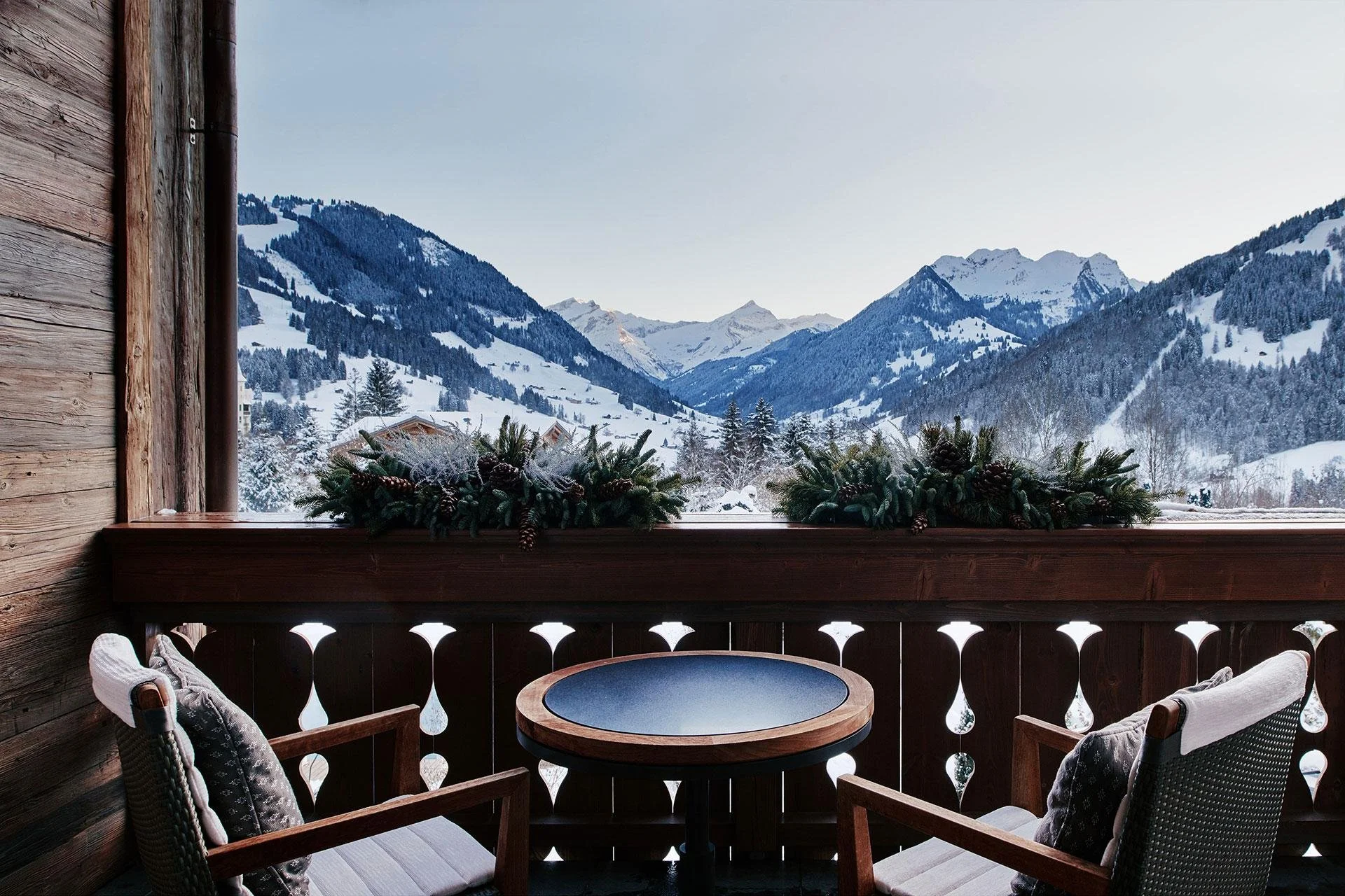 A cozy balcony with wooden railings and chairs overlooking snow-covered mountains and pine forests.