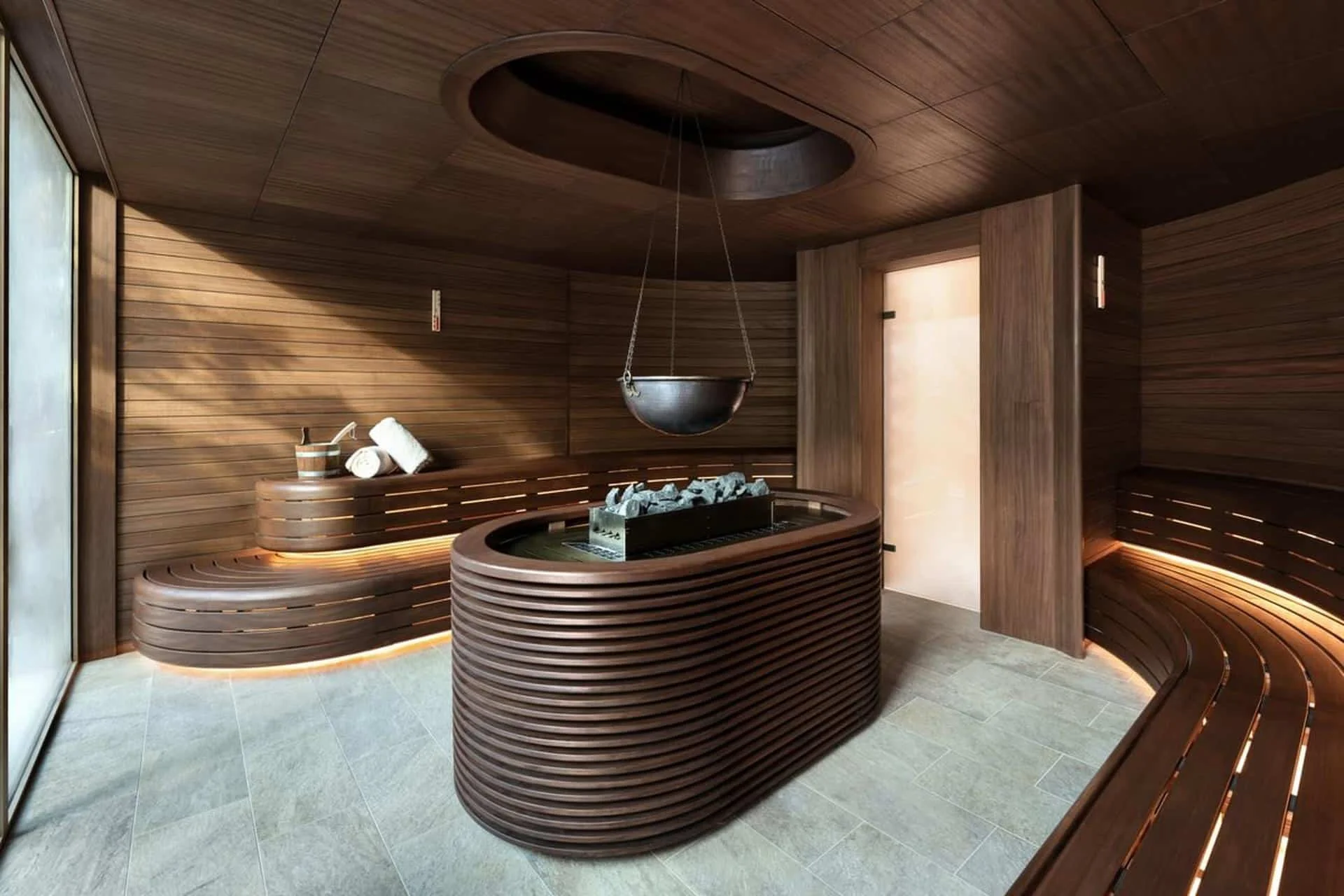 Modern spa room with wooden walls, a curved wooden bench, a suspended metal bowl, and a stone basin filled with ice, designed for a sauna or relaxation.