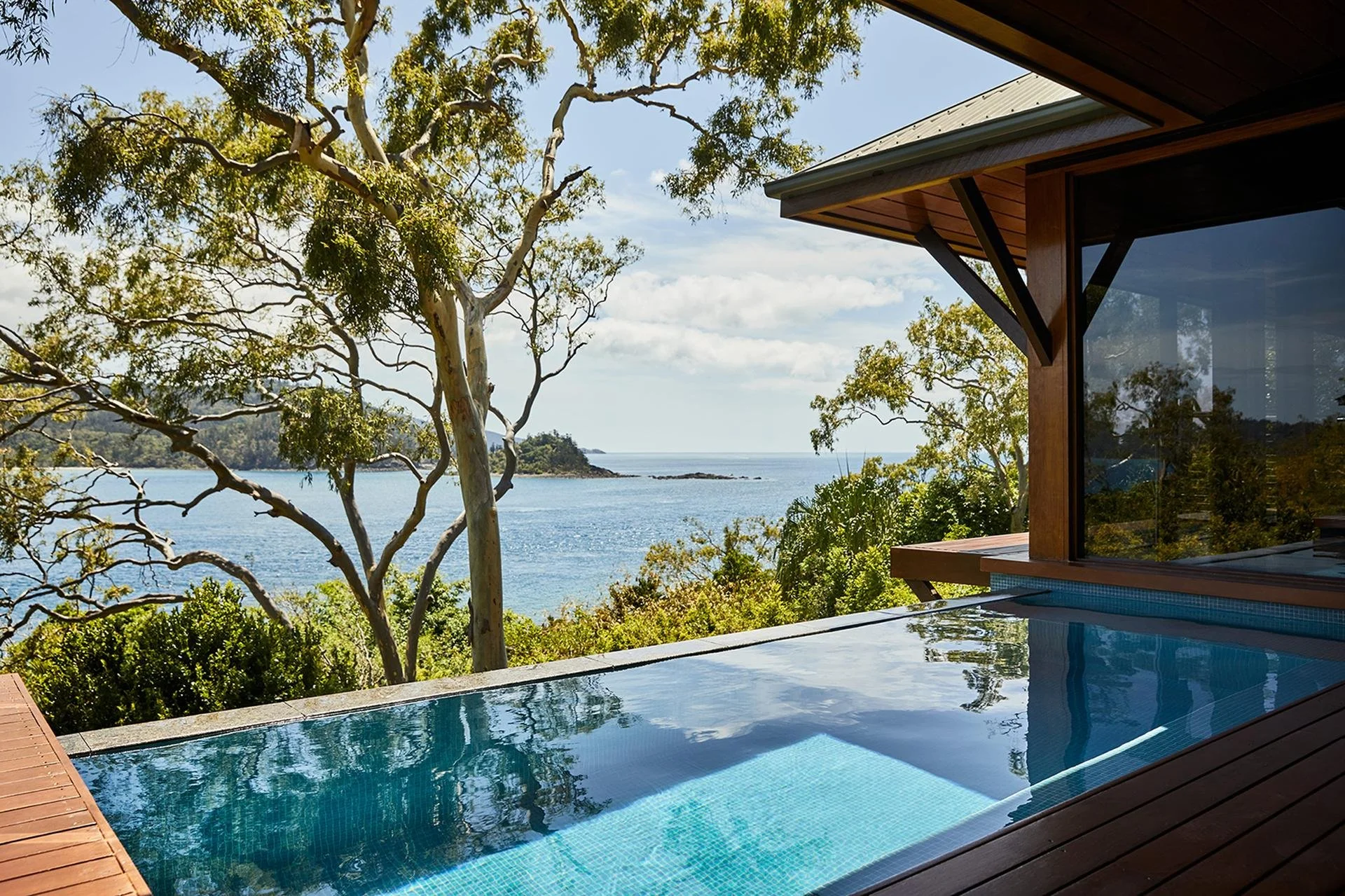 View of a backyard pool with an ocean view, surrounded by trees and a modern house with large windows and a wooden overhang.