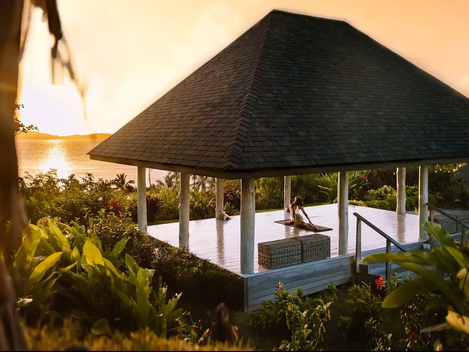 A person practicing yoga on a wooden pavilion with a shingled roof overlooking a sunset by a body of water, surrounded by lush greenery.