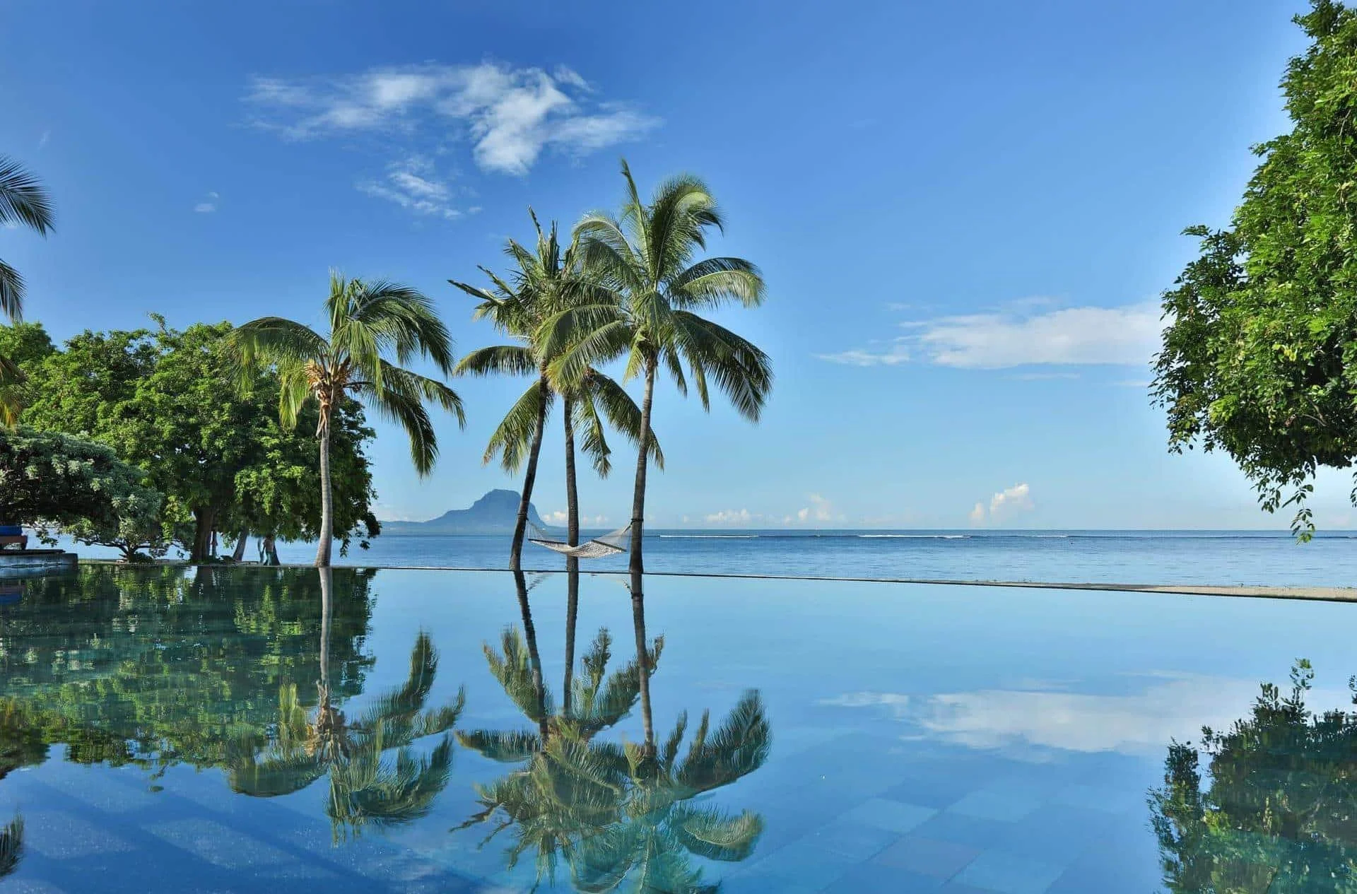 Tropical beach scene with palm trees, a hammock, and a calm pool reflecting the sky and trees, with the ocean and distant island in the background on a sunny day.