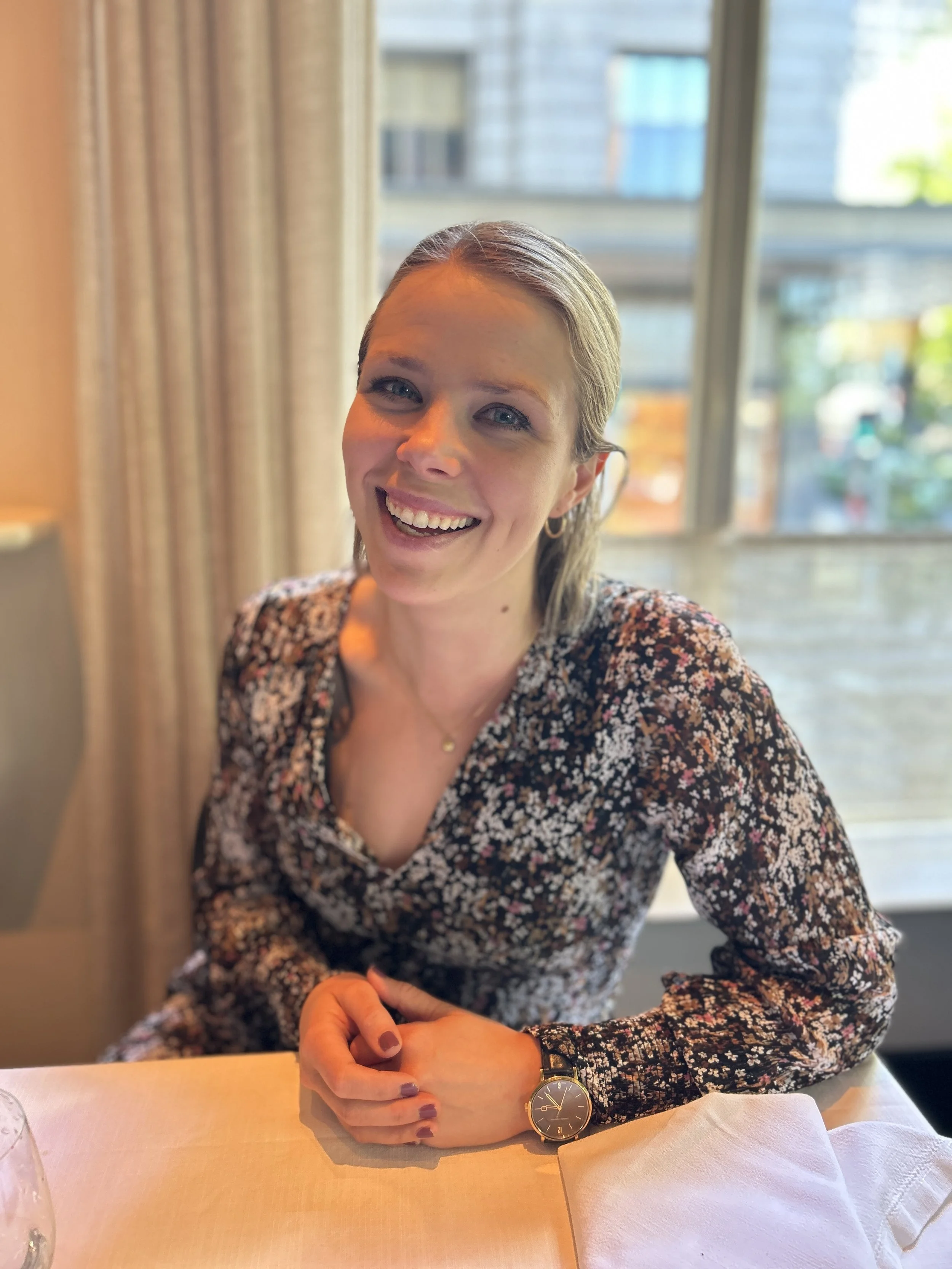 A woman with blonde hair, smiling, sitting at a table in a restaurant or cafe, wearing a floral top, gold watch, and a necklace, with large windows and city buildings in the background.