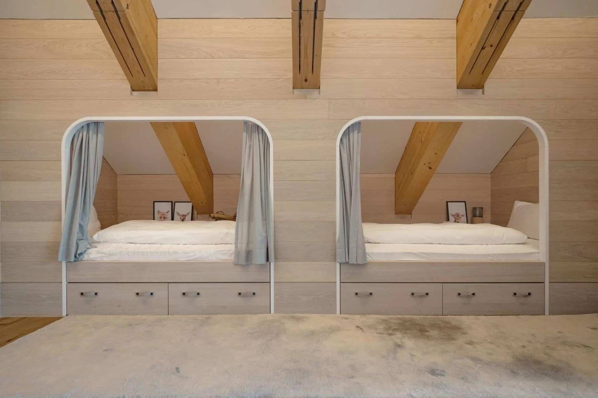 Two beds inside a wooden room with sloped ceilings and exposed beams, with each bed having a white frame, drawers underneath, and curtains for privacy.
