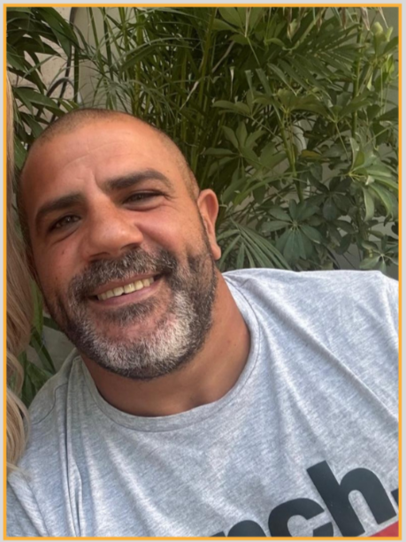 A smiling man with a beard and shaved head, wearing a light gray t-shirt, standing in front of lush green leafy plants.
