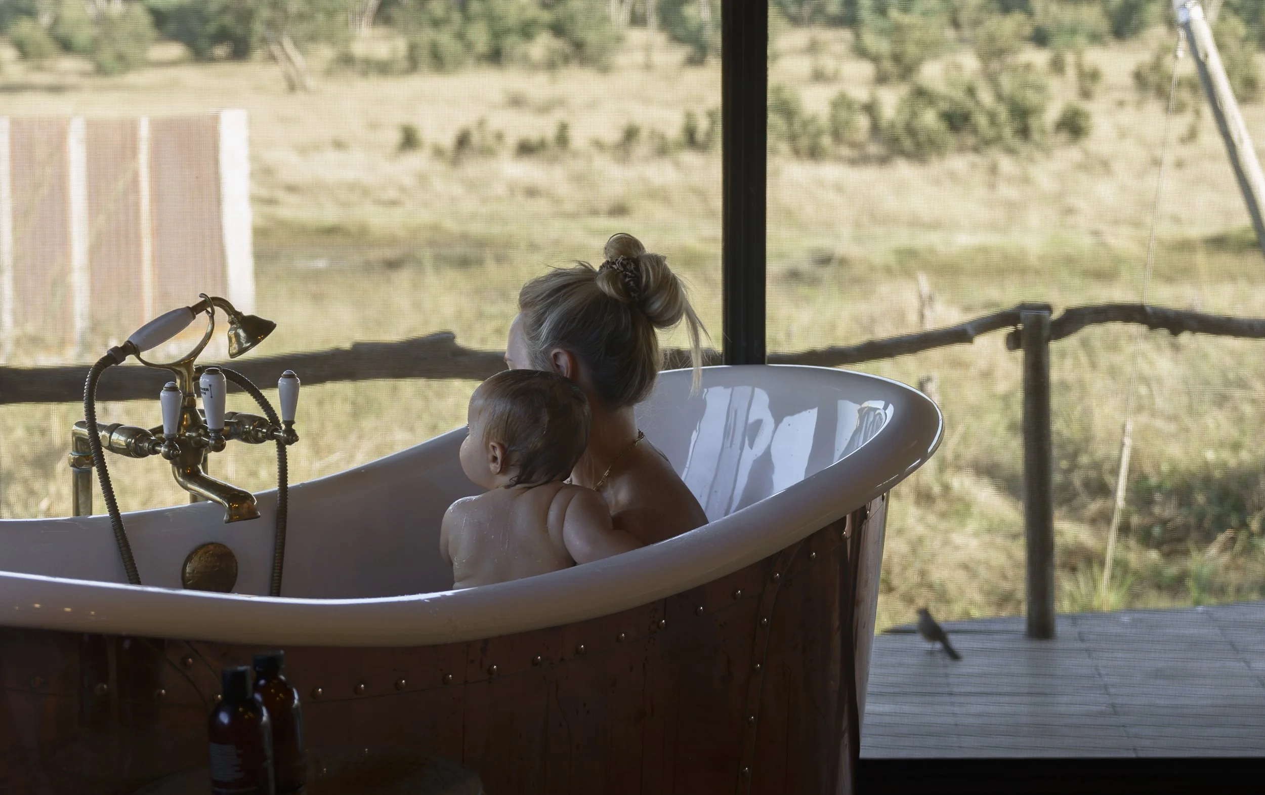 Mother bathing young child in a freestanding tub overlooking the bush through a mesh window
