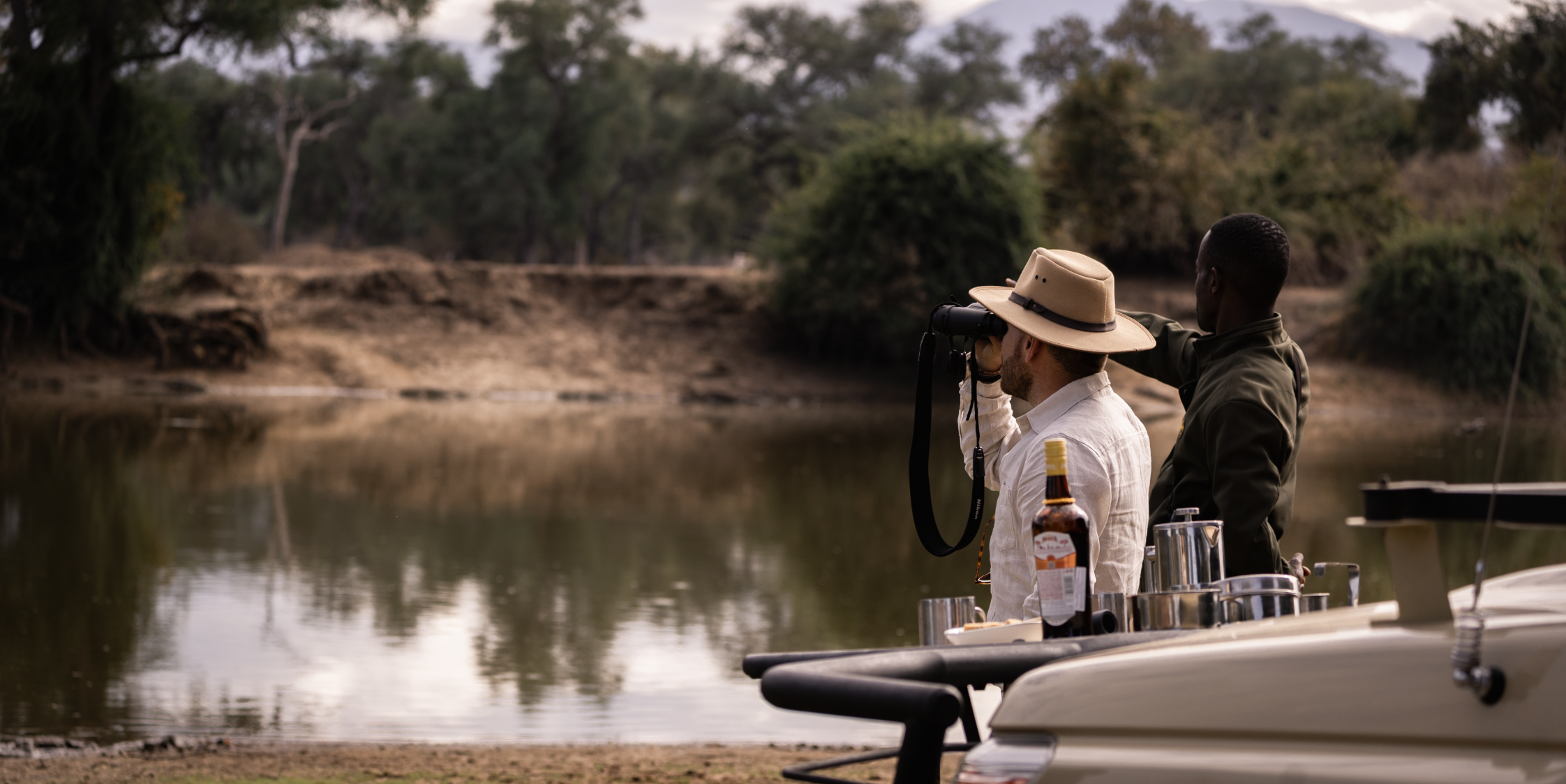 Two men on a boat, one using binoculars, overlooking a river with trees in the background, during sunset.