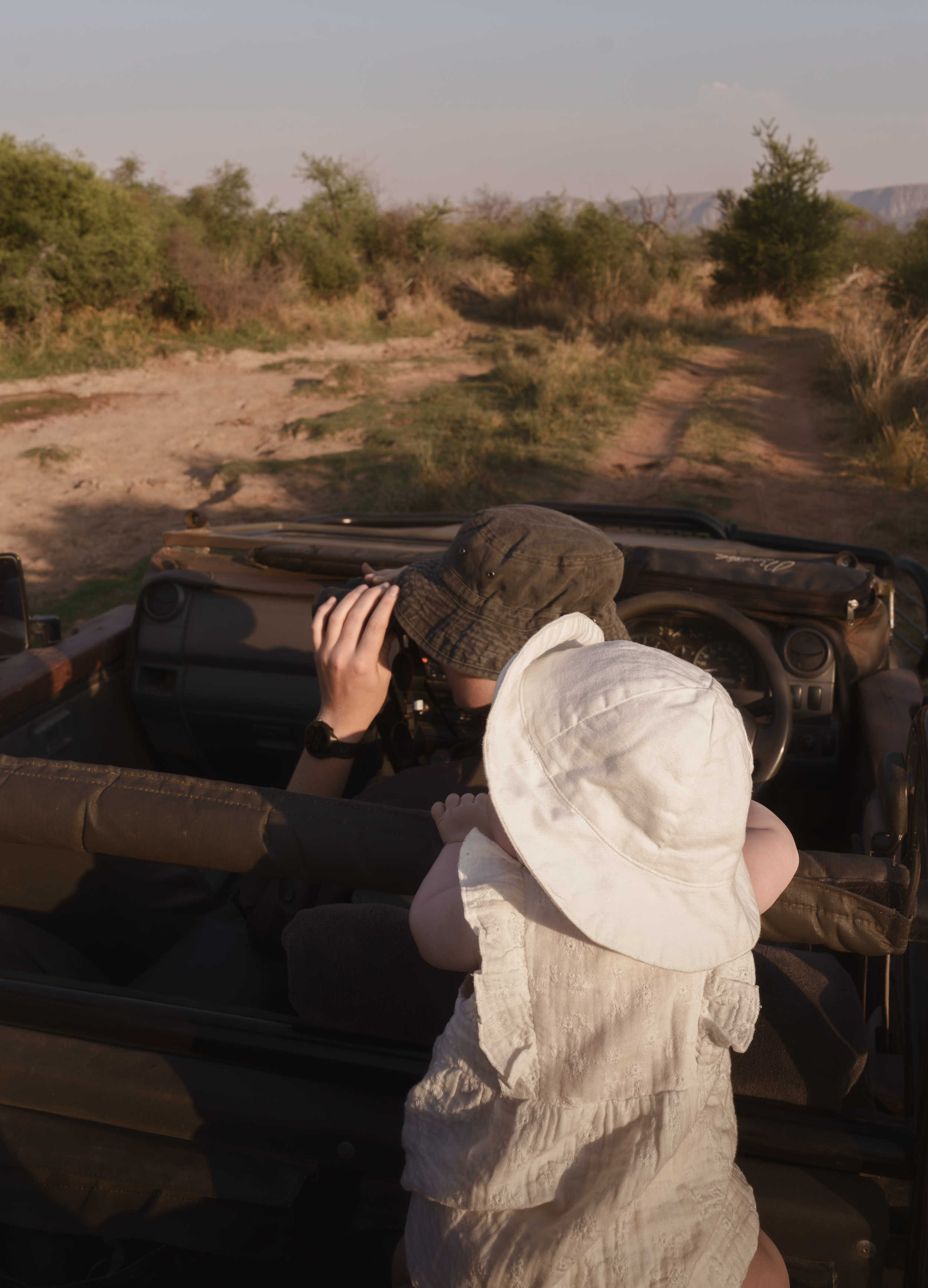 Baby in a sun hat standing on a safari vehicle seat beside a parent watching through binoculars