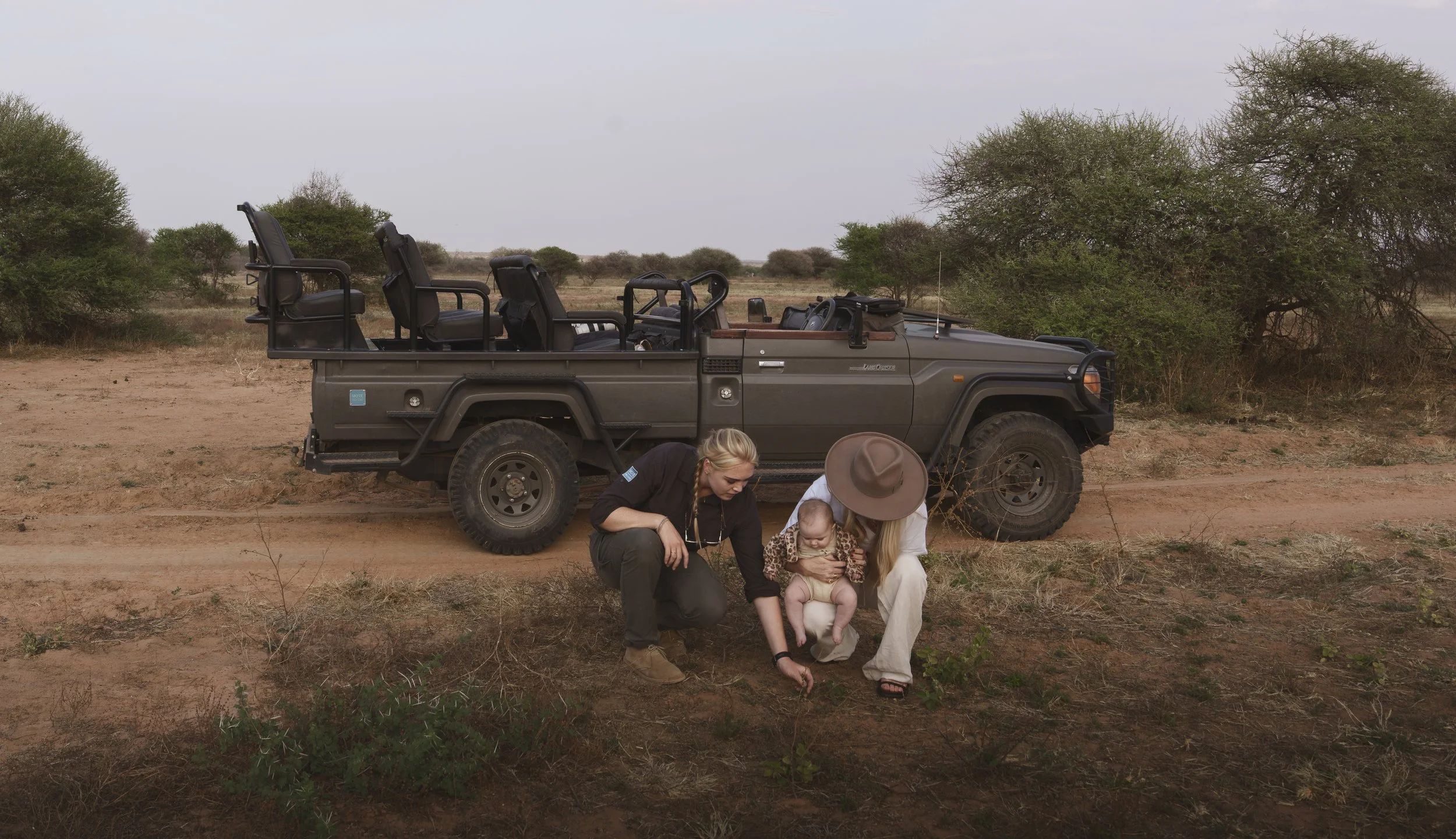 guide crouching beside a game drive vehicle showing a baby a plant, held by her mother